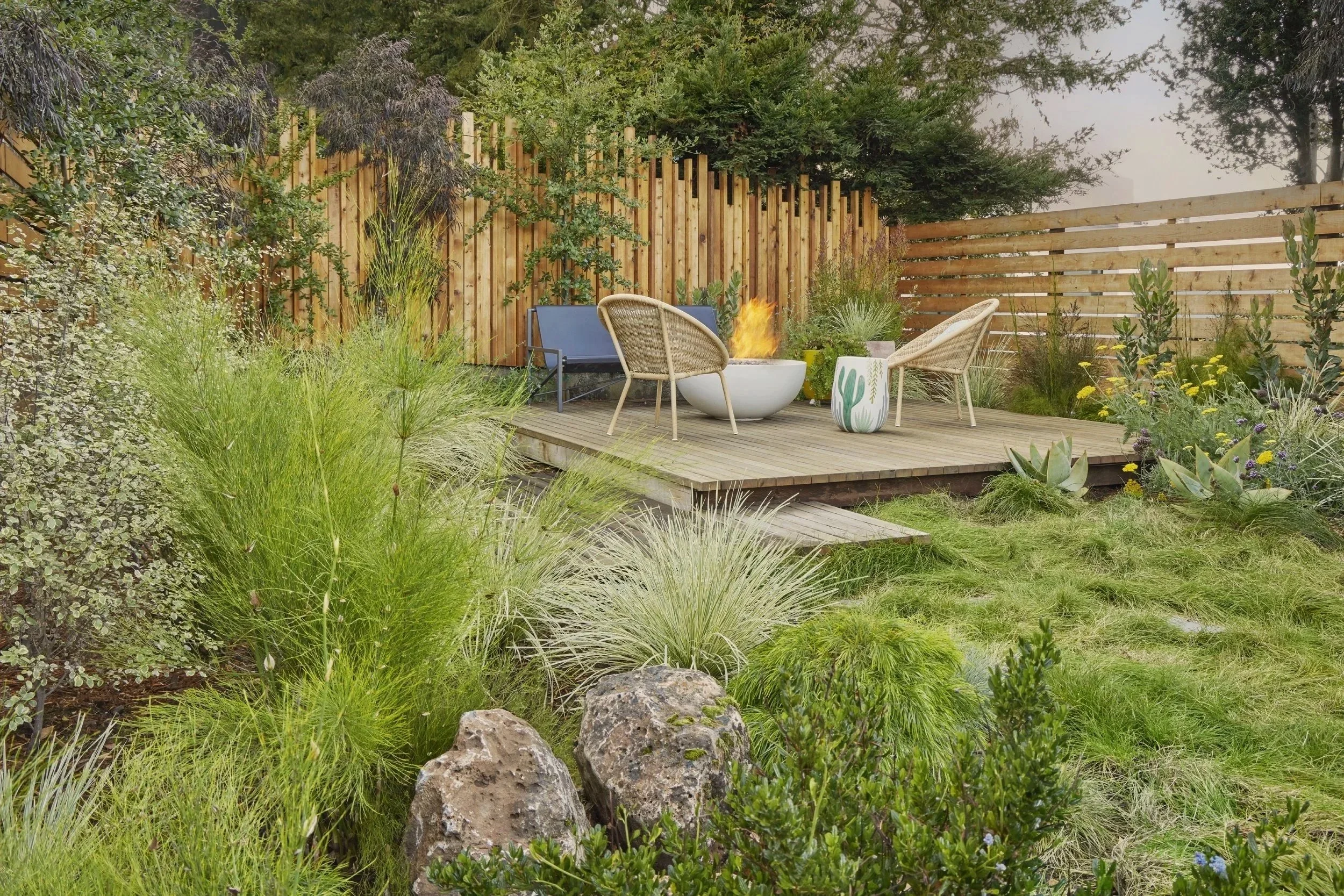 A cozy backyard patio with a fire pit, cushioned chairs, and a variety of lush green plants and ornamental grasses, enclosed by wooden fencing.