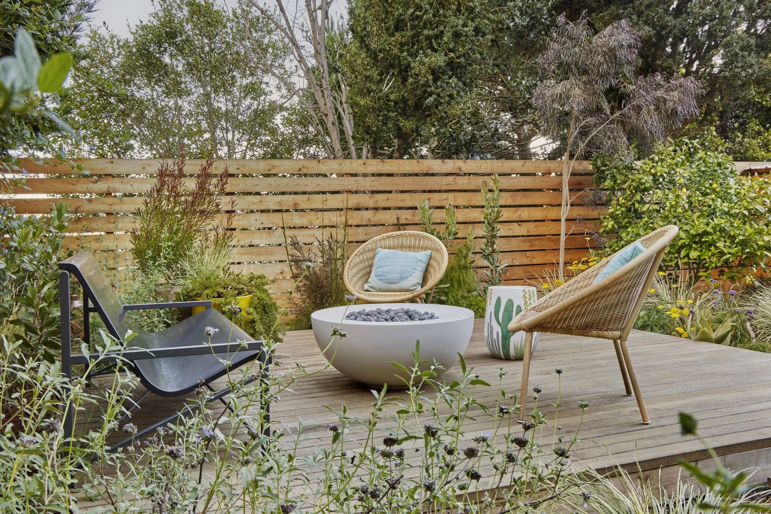 A cozy outdoor patio with a wooden deck, surrounded by lush greenery, including shrubs, bushes, and trees. The patio features wicker chairs with pillows, a modern white fire pit with black stones, and a painted plant pot, creating a relaxing garden s