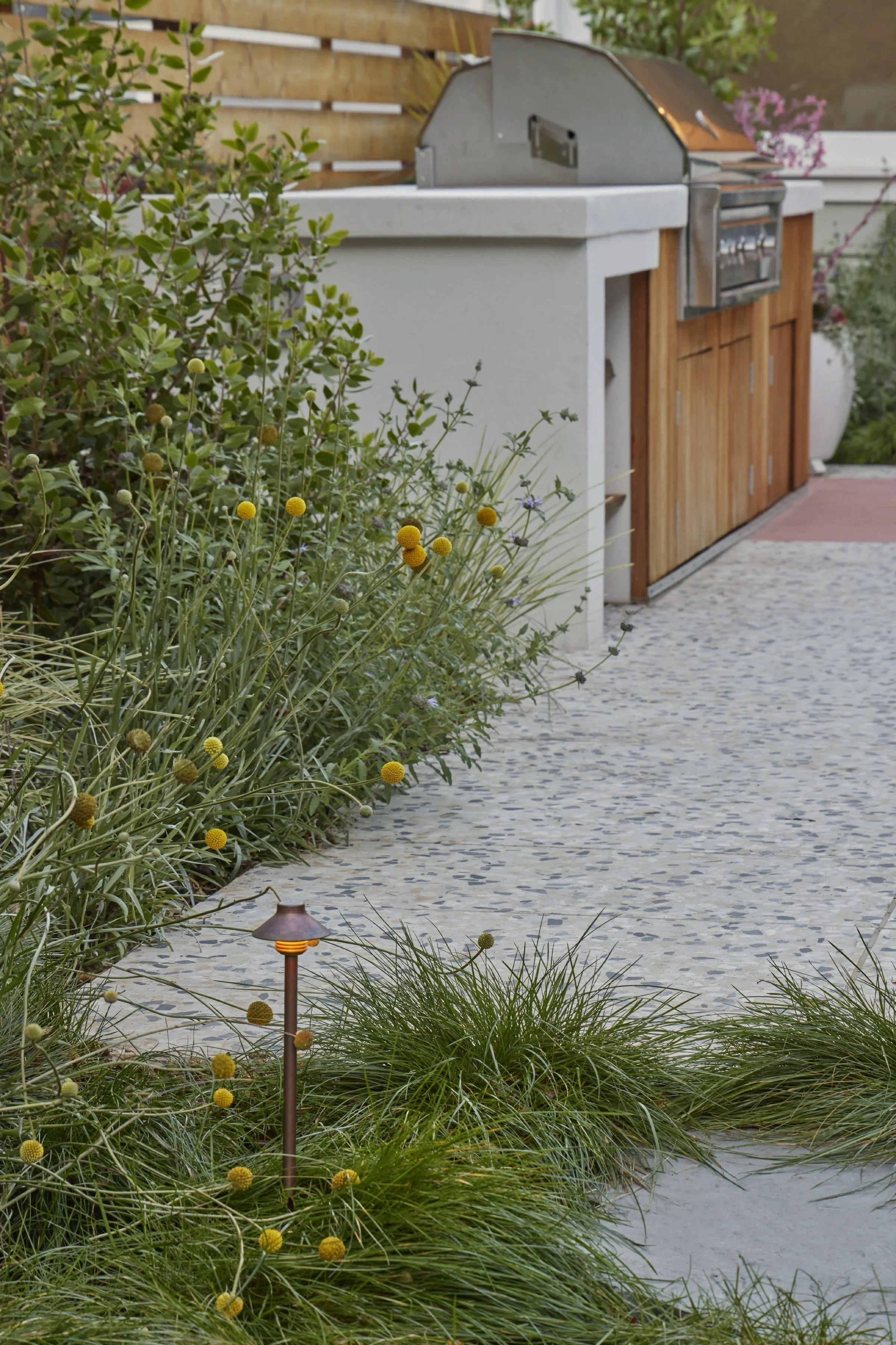Outdoor patio with a built-in grill, surrounded by lush green plants and flowering bushes, and a pebble pathway.