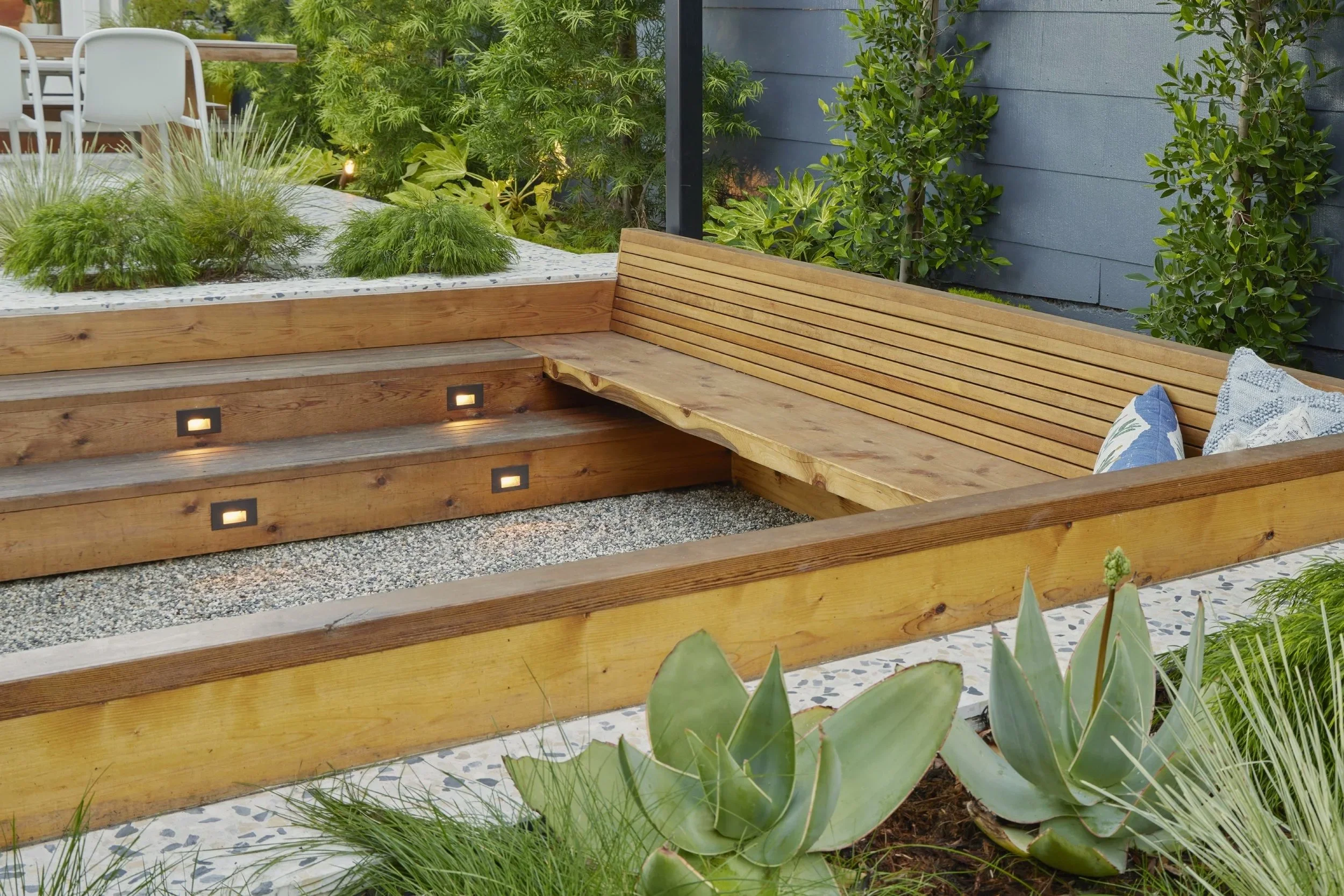 Outdoor wooden seating area with built-in bench, small decorative lights, plants, and pillows, next to a garden with green shrubs and succulents.