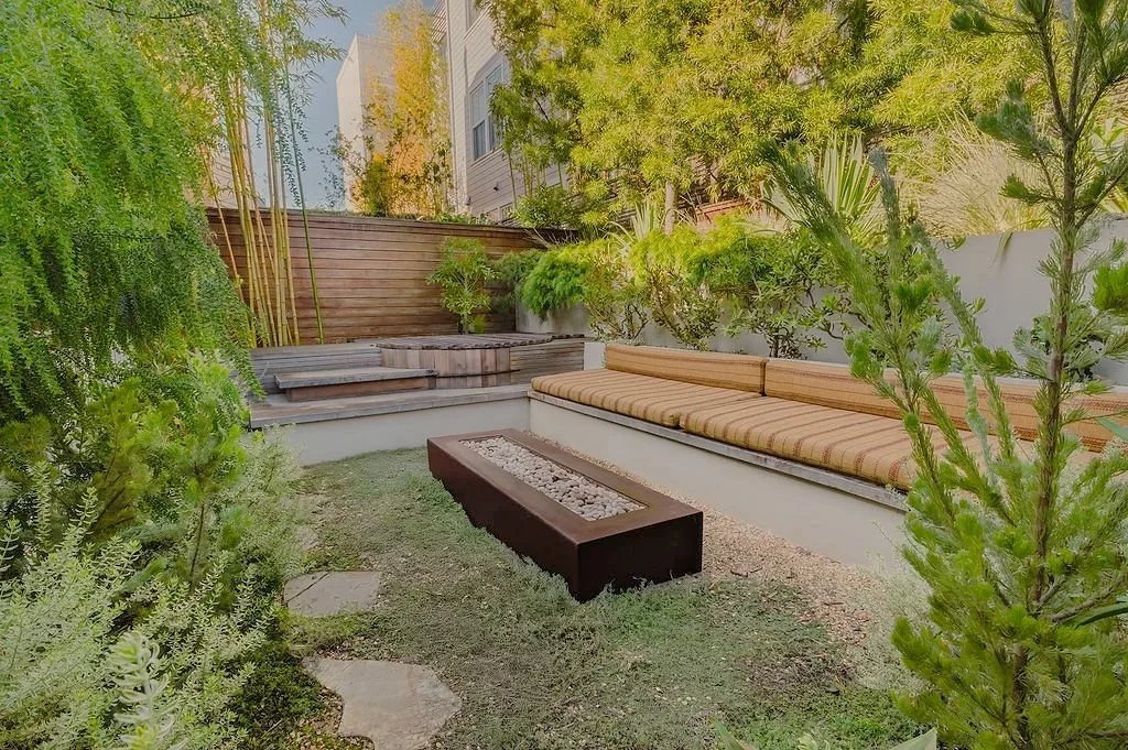 Small backyard with built-in bench seating, a rectangular fire pit, and a variety of green plants and trees.