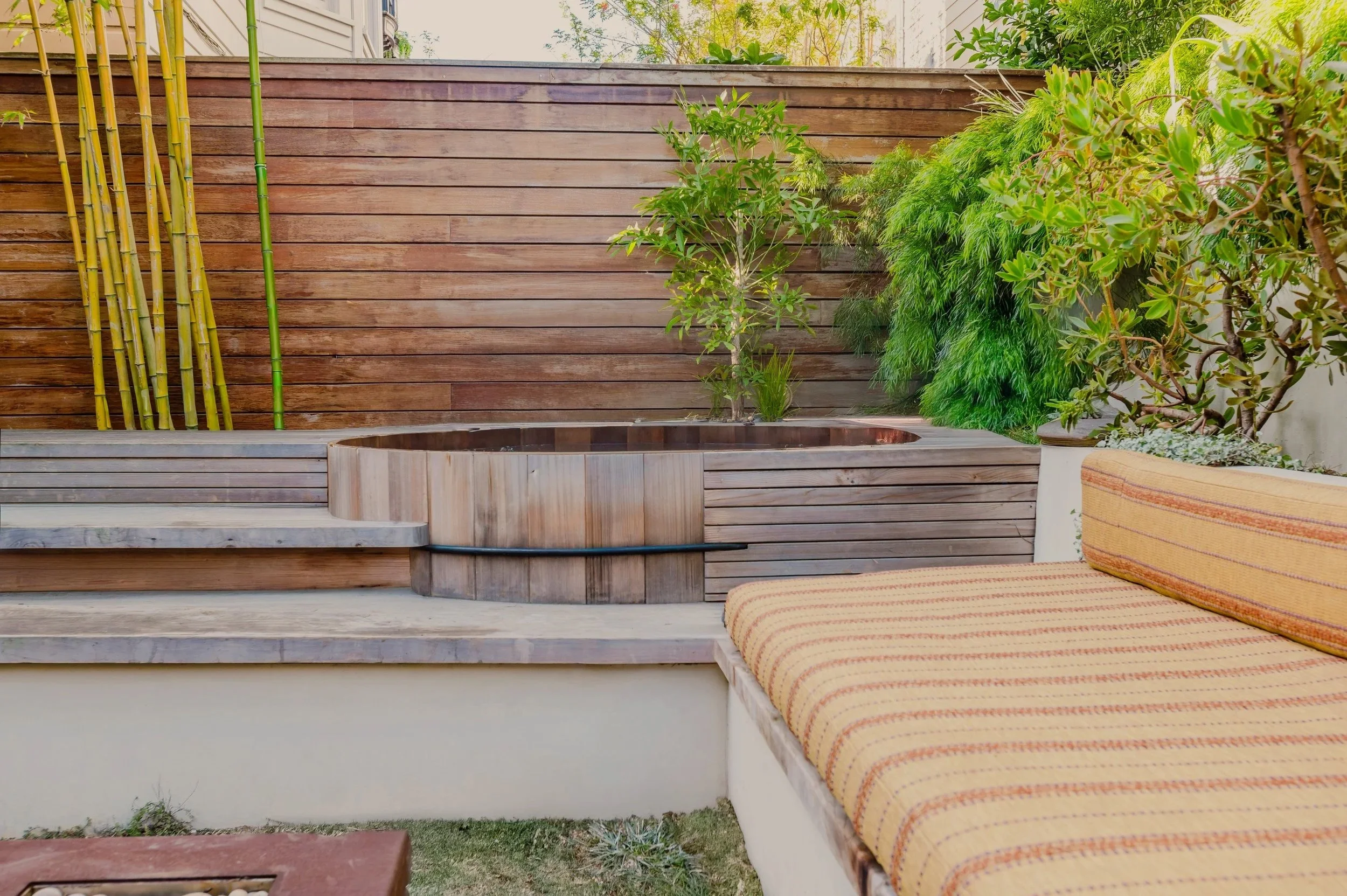 An outdoor patio featuring wooden decking, a built-in hot tub, green plants, bamboo stalks, and a cushioned seating area with striped fabric.