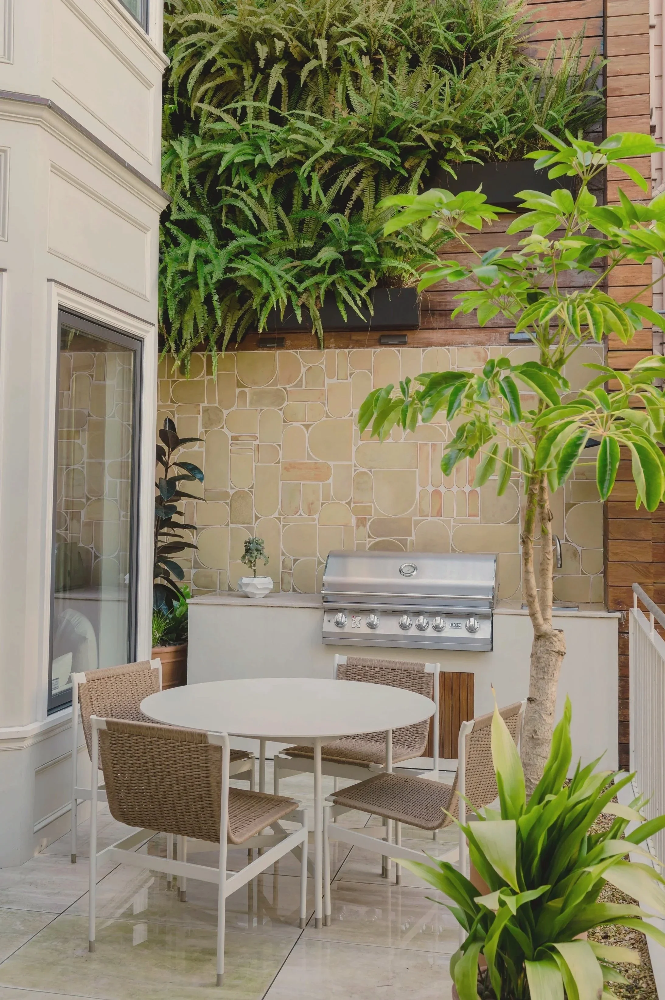 Outdoor patio with a white round table and six chairs, a built-in grill, tropical plants including a tree, potted fern, and leafy green plant, beige tiled wall, wooden paneling, and a window.