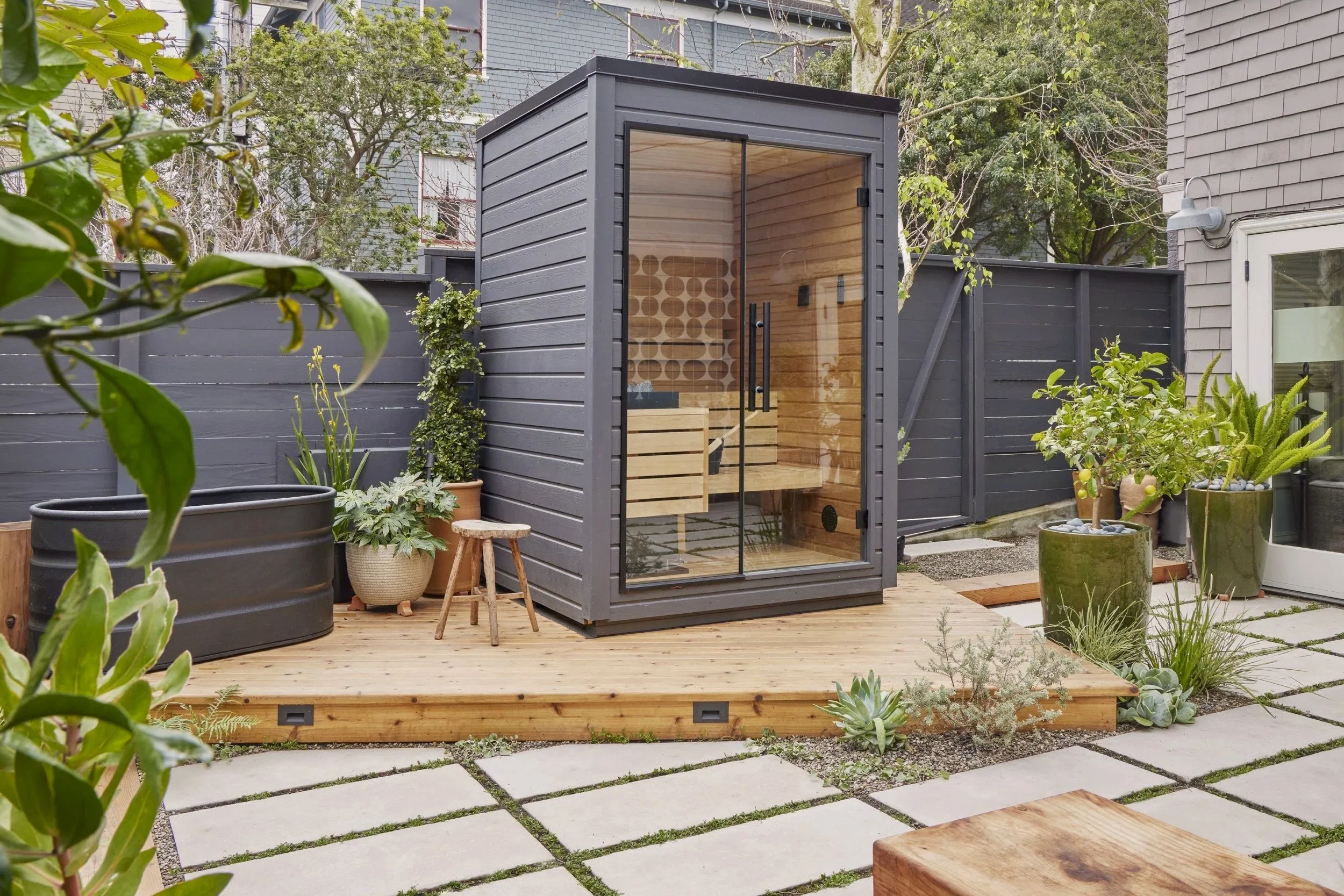 A backyard patio with a wooden decking area featuring a gray outdoor sauna with glass door, surrounded by potted plants and greenery, and a paved stone pathway.
