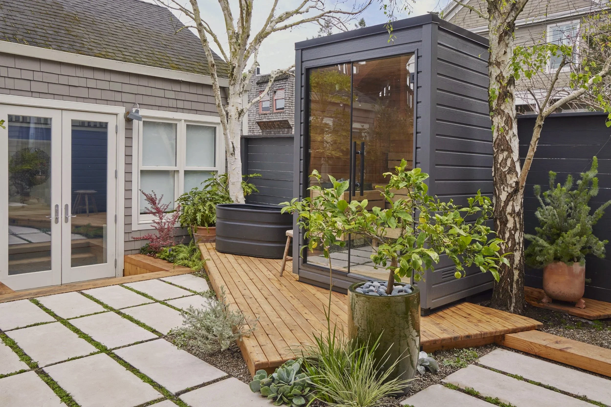 Small backyard with a wooden deck, potted plants, and a black garden shed with large glass doors. A tree with peeling bark and other greenery are visible.