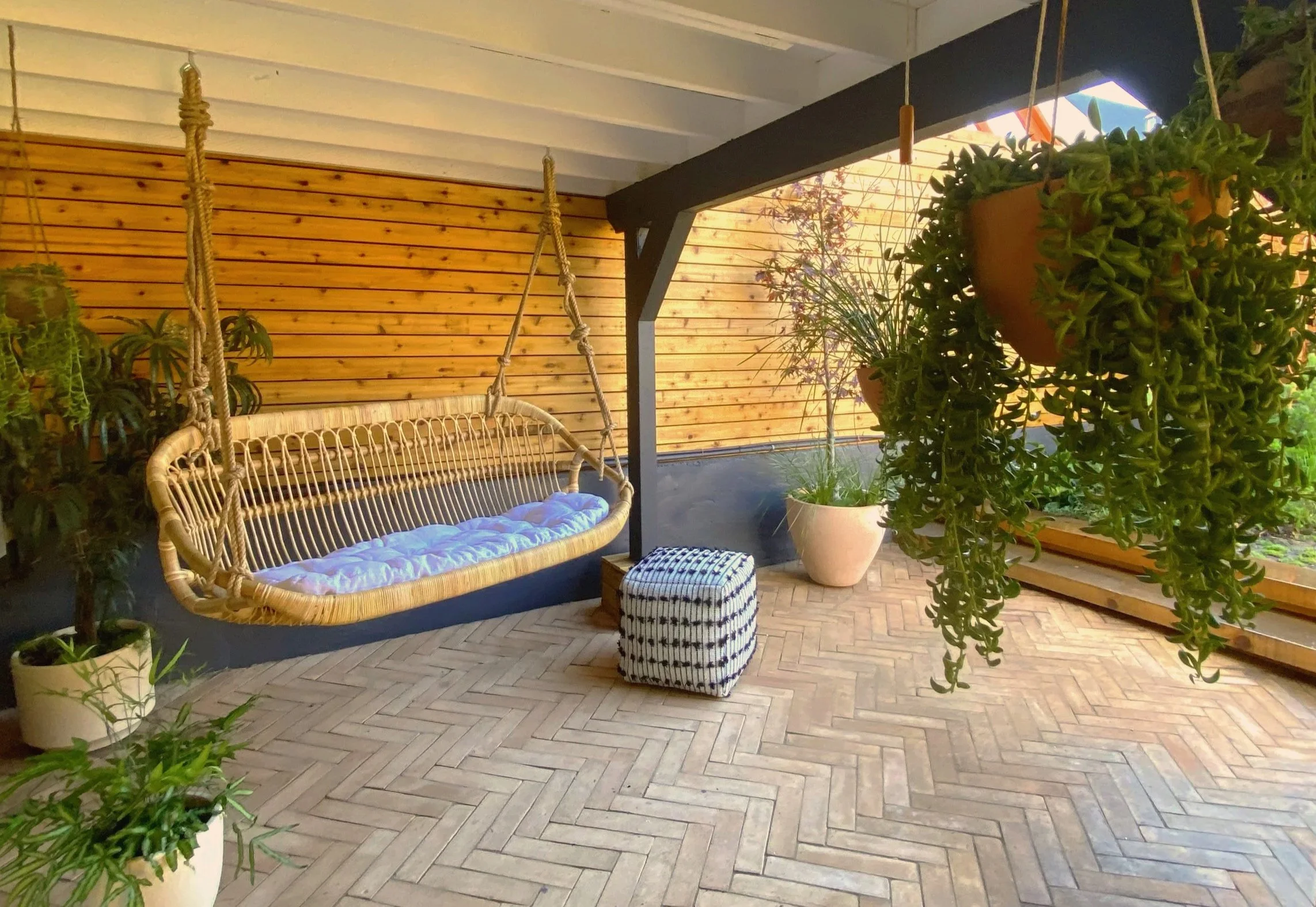Patio area with a wooden slat wall, hanging wicker bench with blue cushion, potted plants, and hanging planters, with a black-painted wooden support beam and a herringbone-patterned brick floor.