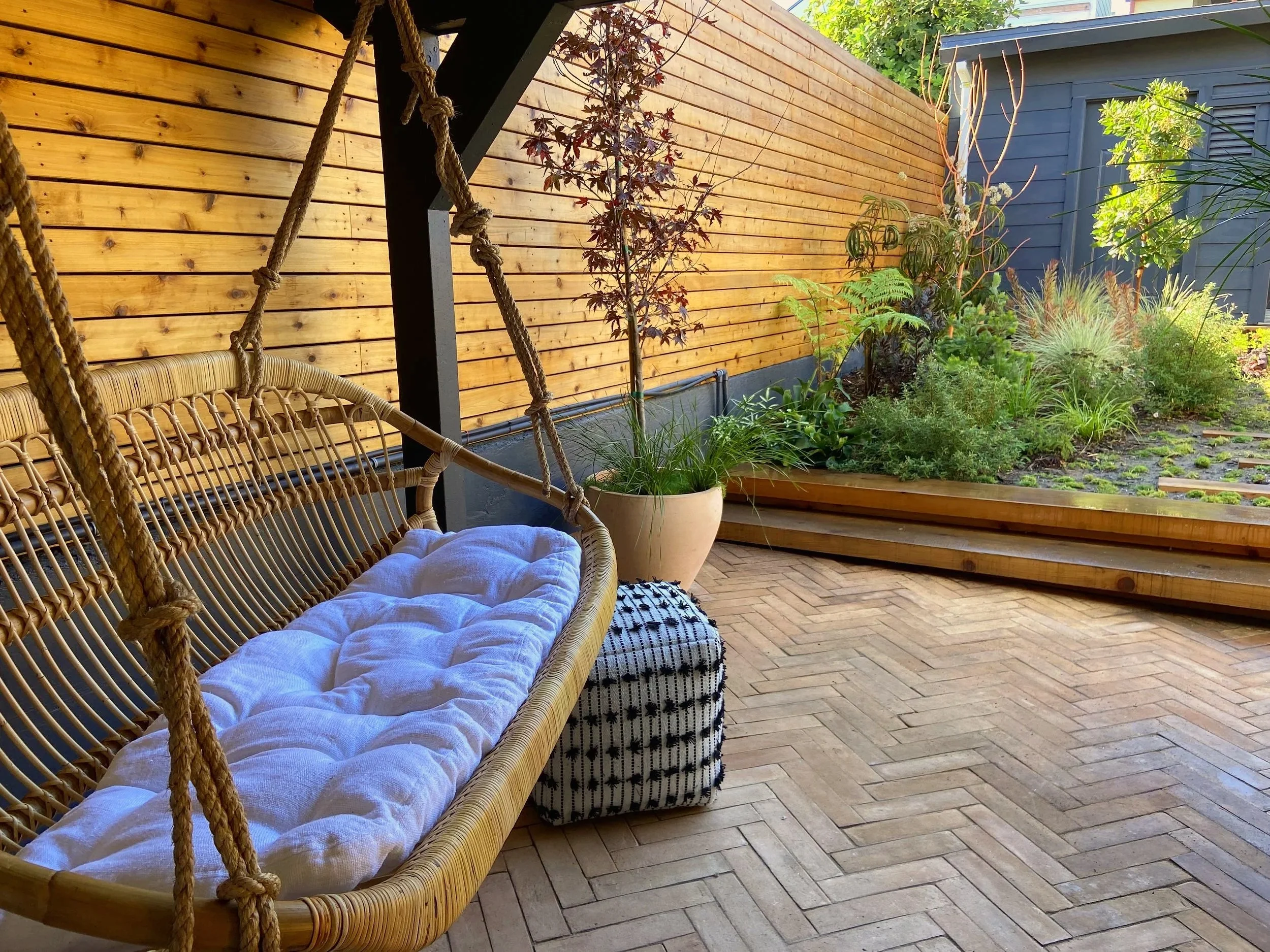 Outdoor patio with a wooden hanging swing, a large potted plant, and a garden with various green plants behind a wooden fence.
