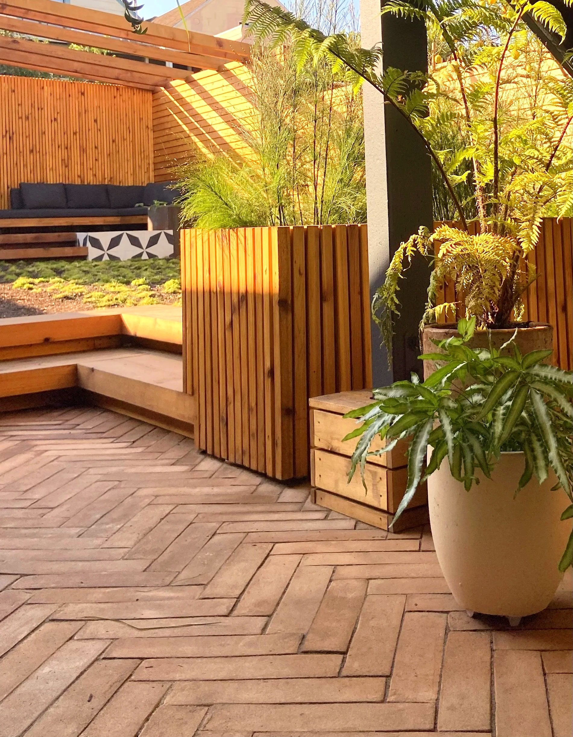 An outdoor patio area with wooden flooring in a herringbone pattern, potted plants including ferns and leafy plants, wooden benches, and a sitting area with black cushions. Sunlight creates shadows through a wooden pergola.