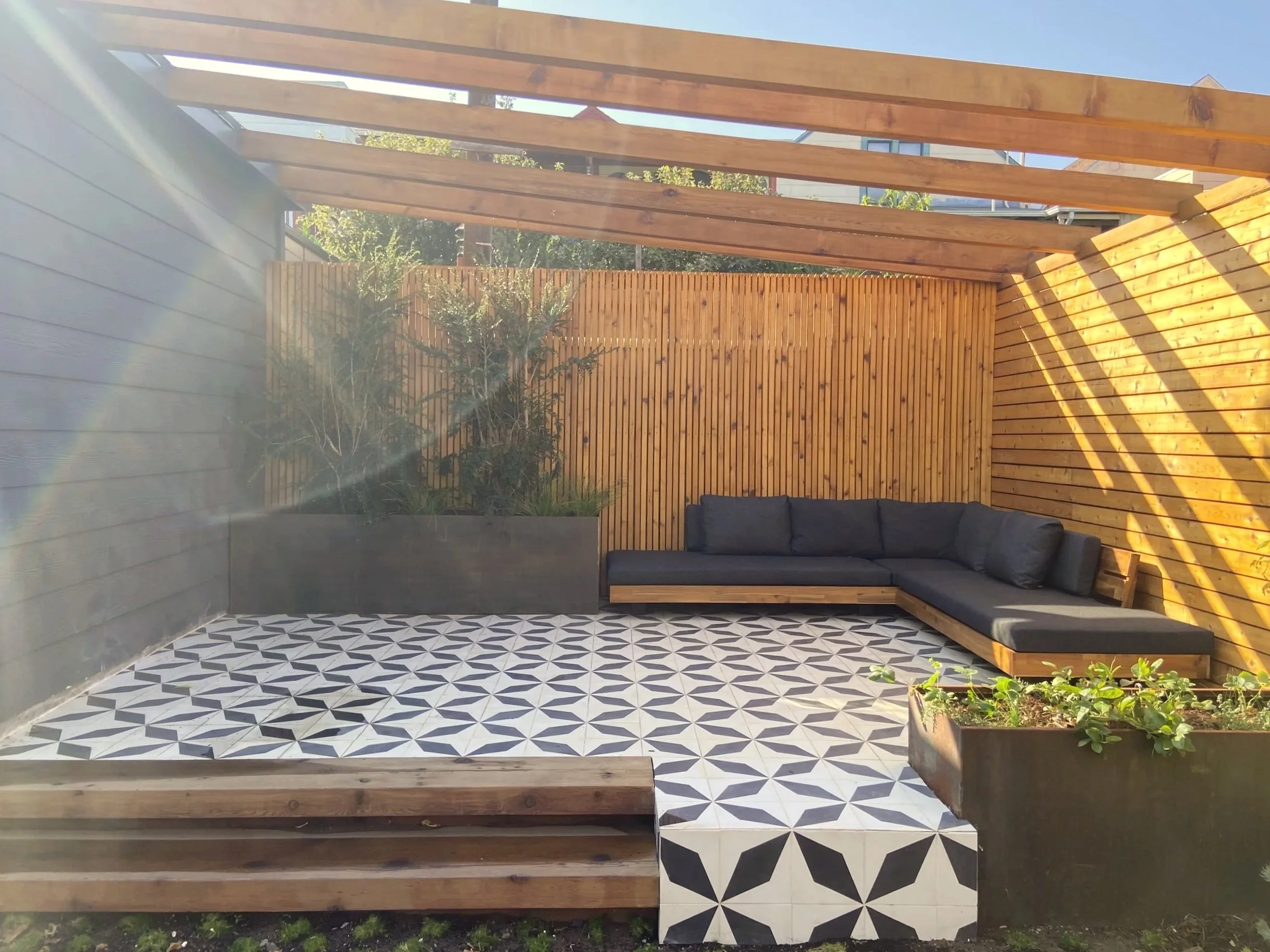A modern outdoor patio with black and white geometric patterned tiles, enclosed by wooden fences. There is a black L-shaped outdoor sofa with cushions on the right side and a large planter with green plants on the right front. A wooden bench is at th