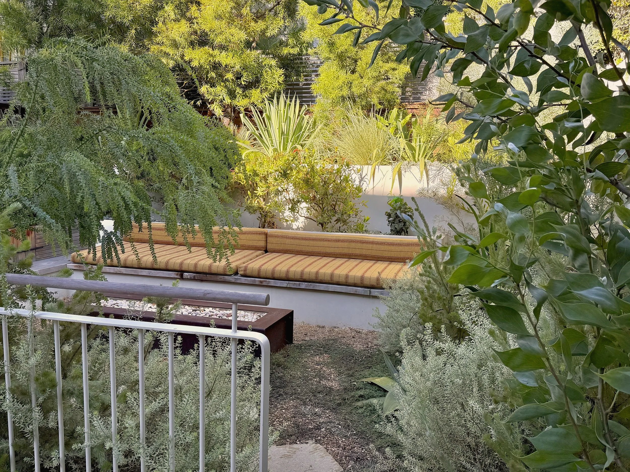 A lush garden with various green plants and shrubs surrounding a beige or tan cushioned outdoor bench under the trees.