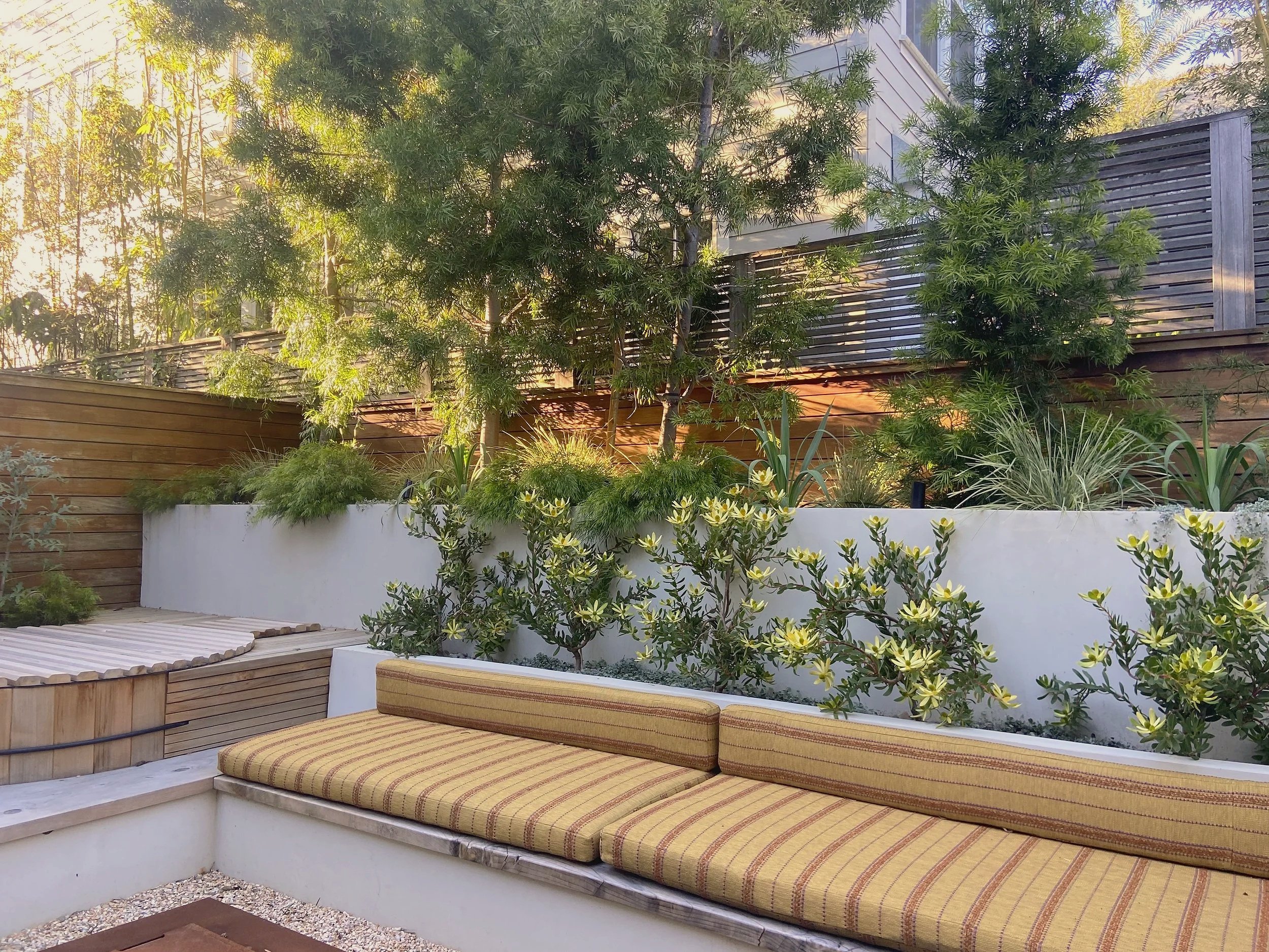 Outdoor seating area with striped cushions, surrounded by lush green plants and trees, with a wooden fence and modern building in the background.