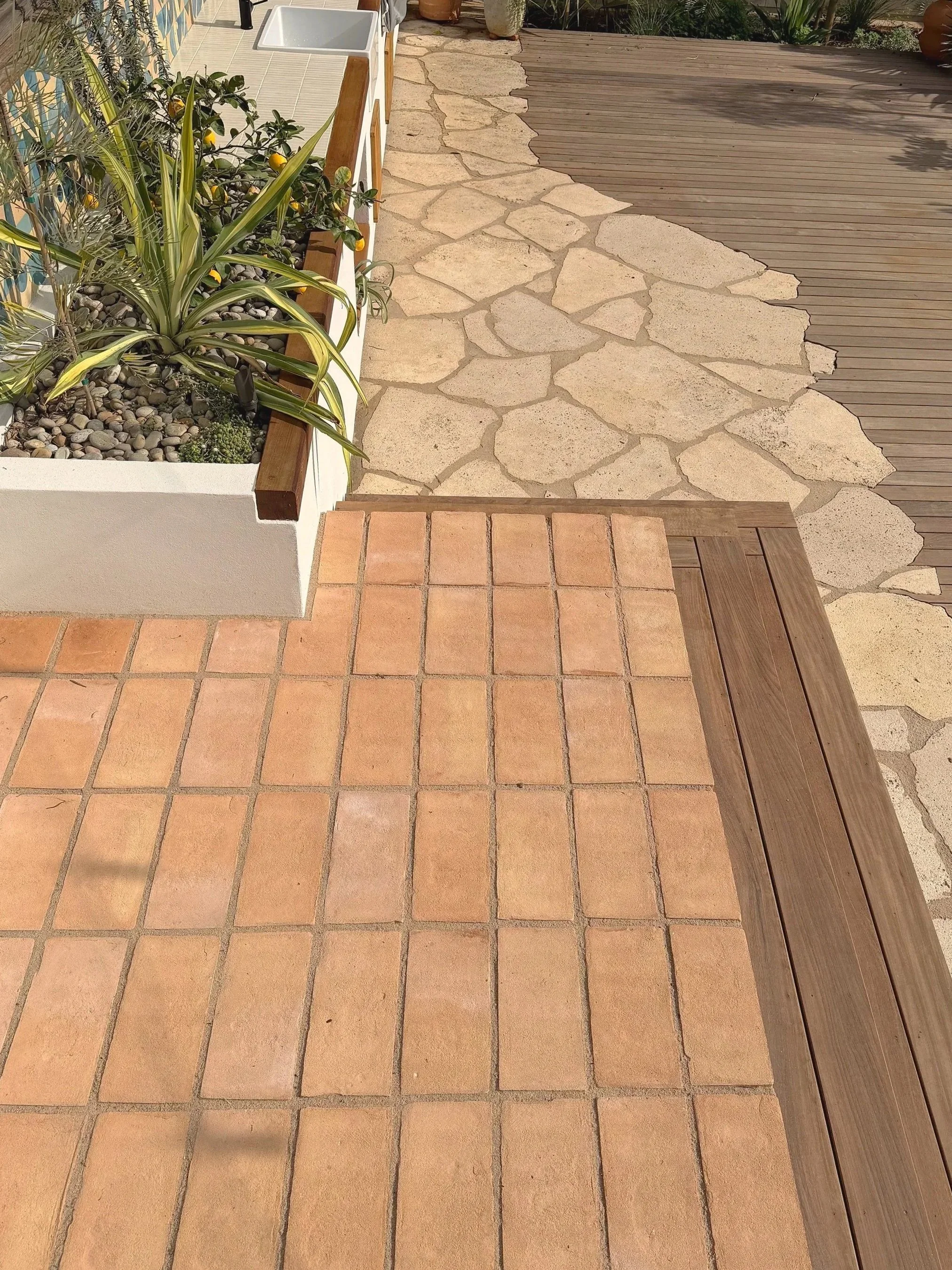 Terracotta tiled patio, transitioning to a stone pathway with irregularly shaped light-colored stones, and a wooden deck on the right side.