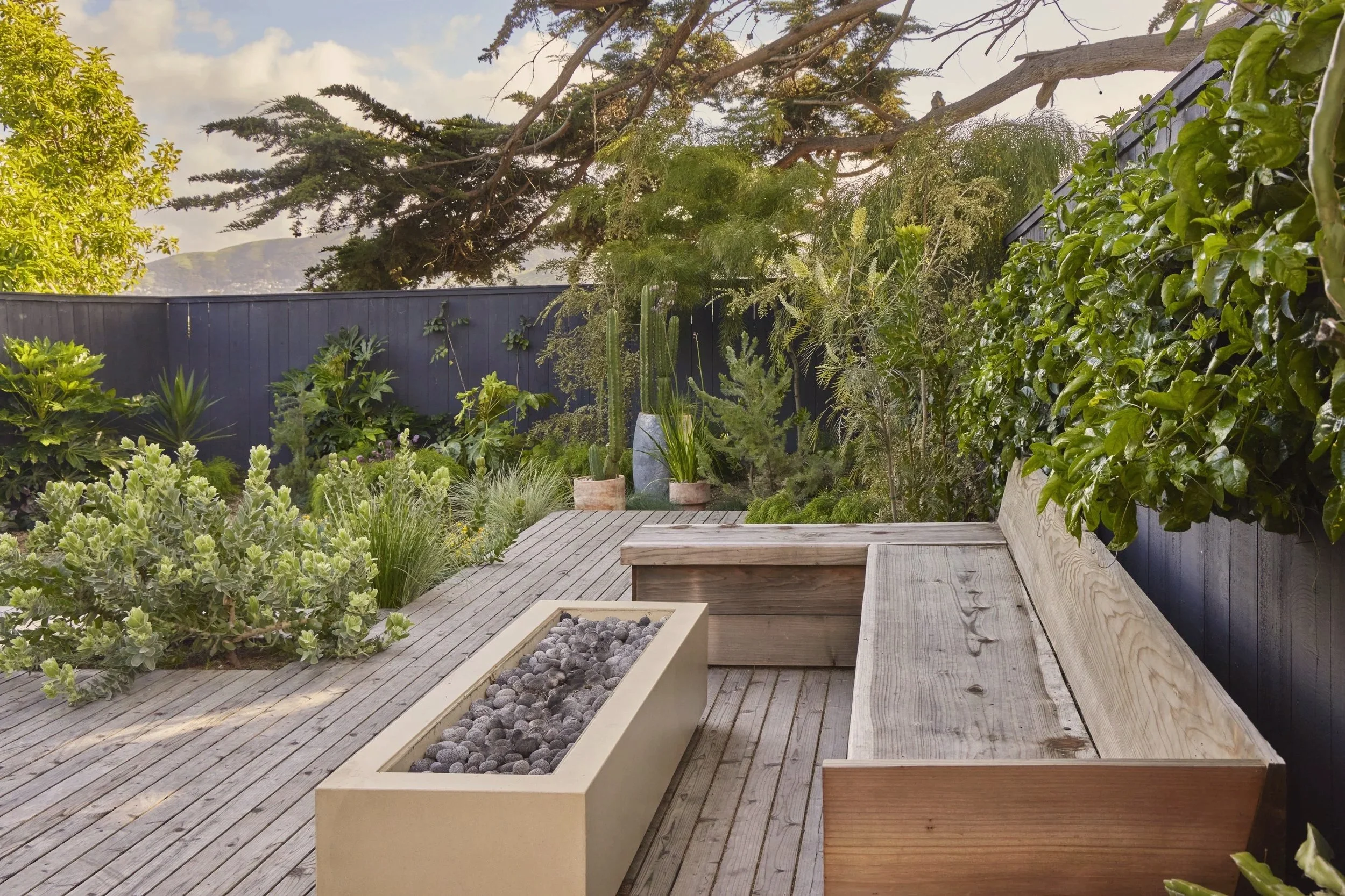 A cozy outdoor patio with a wooden deck, a built-in wooden bench, a rectangular fire pit filled with decorative stones, and lush green plants and trees surrounding the area.