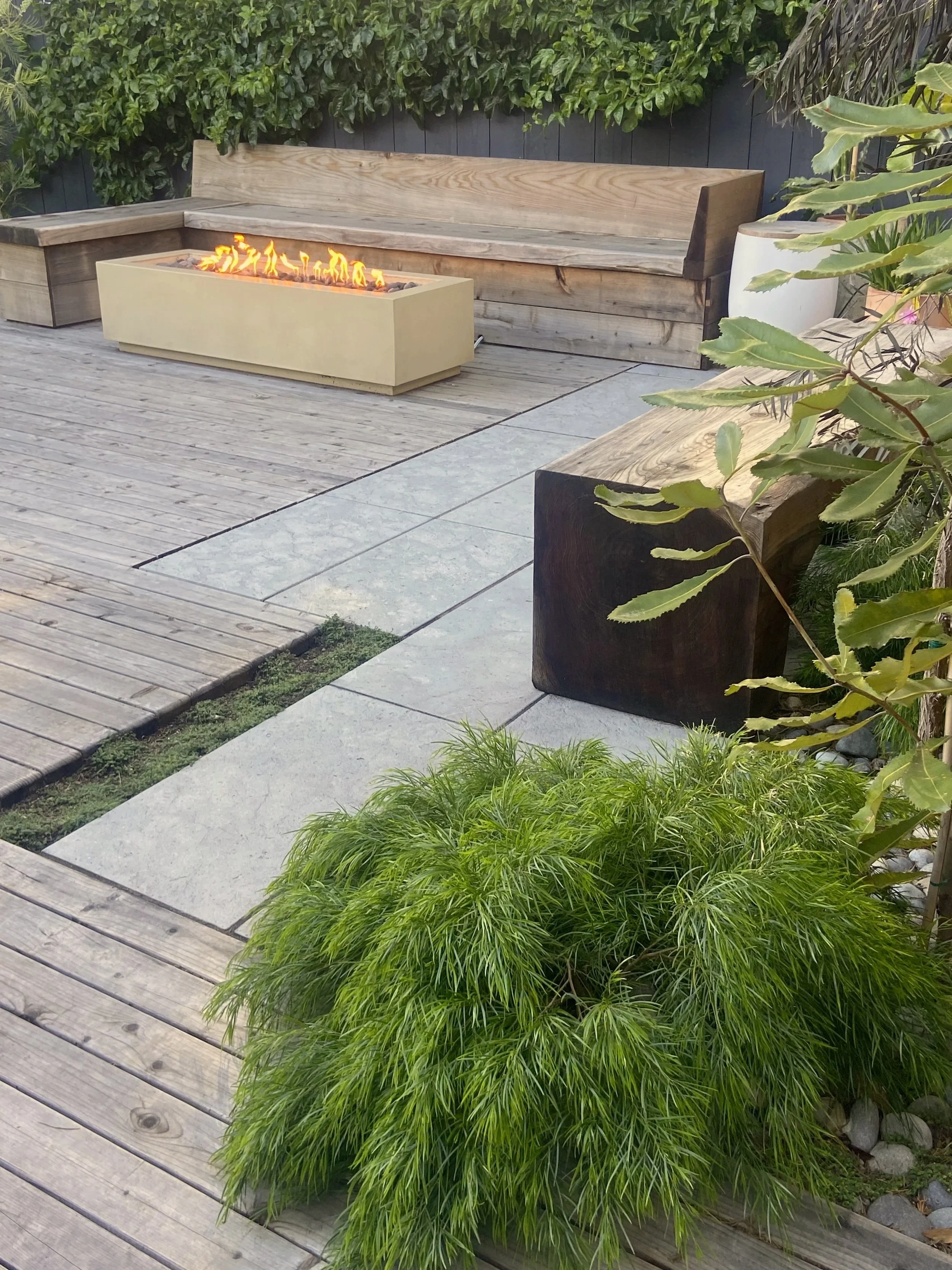 Outdoor patio with wooden decking and concrete tiles, a modern rectangular fire pit with flames, wooden benches, green plants and trees along the border.