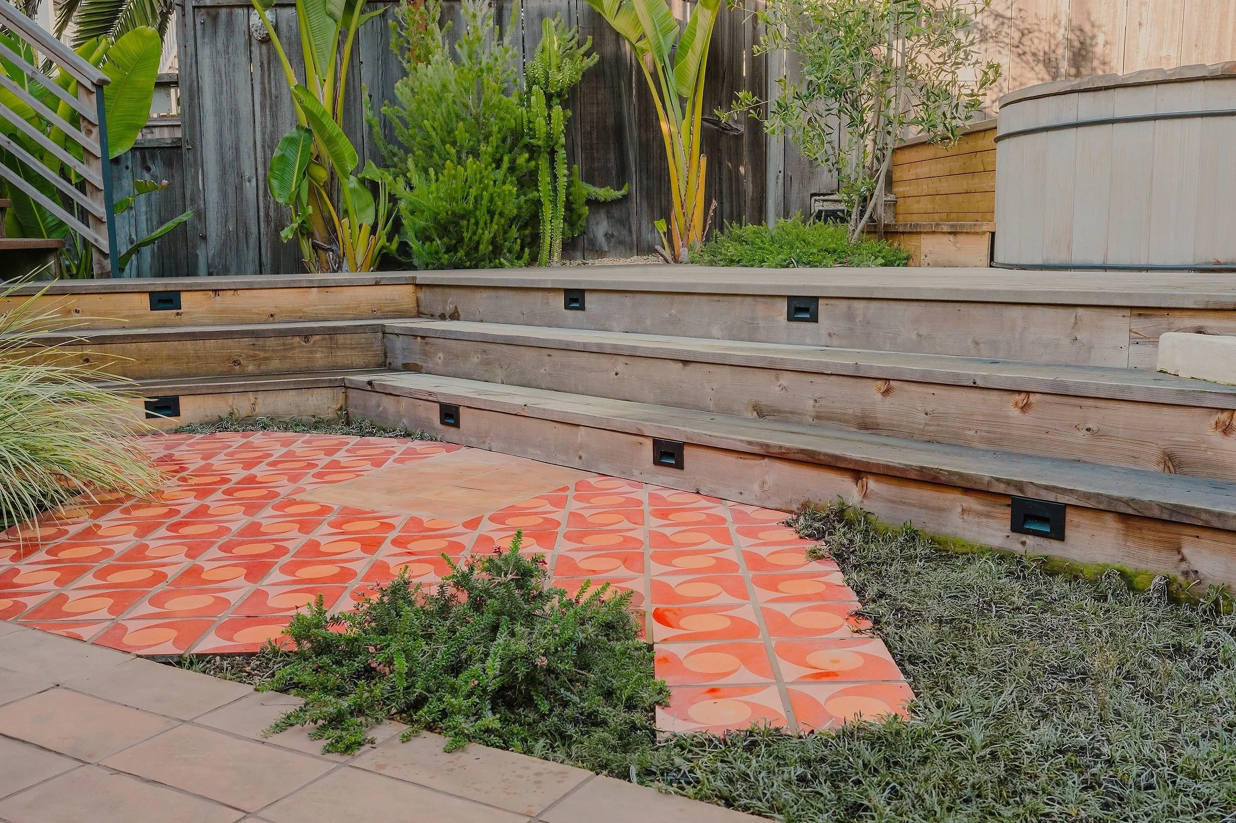 Backyard garden area with orange patterned tiles, wooden stairs, and various green plants including cacti and shrubs, surrounded by a wooden fence.