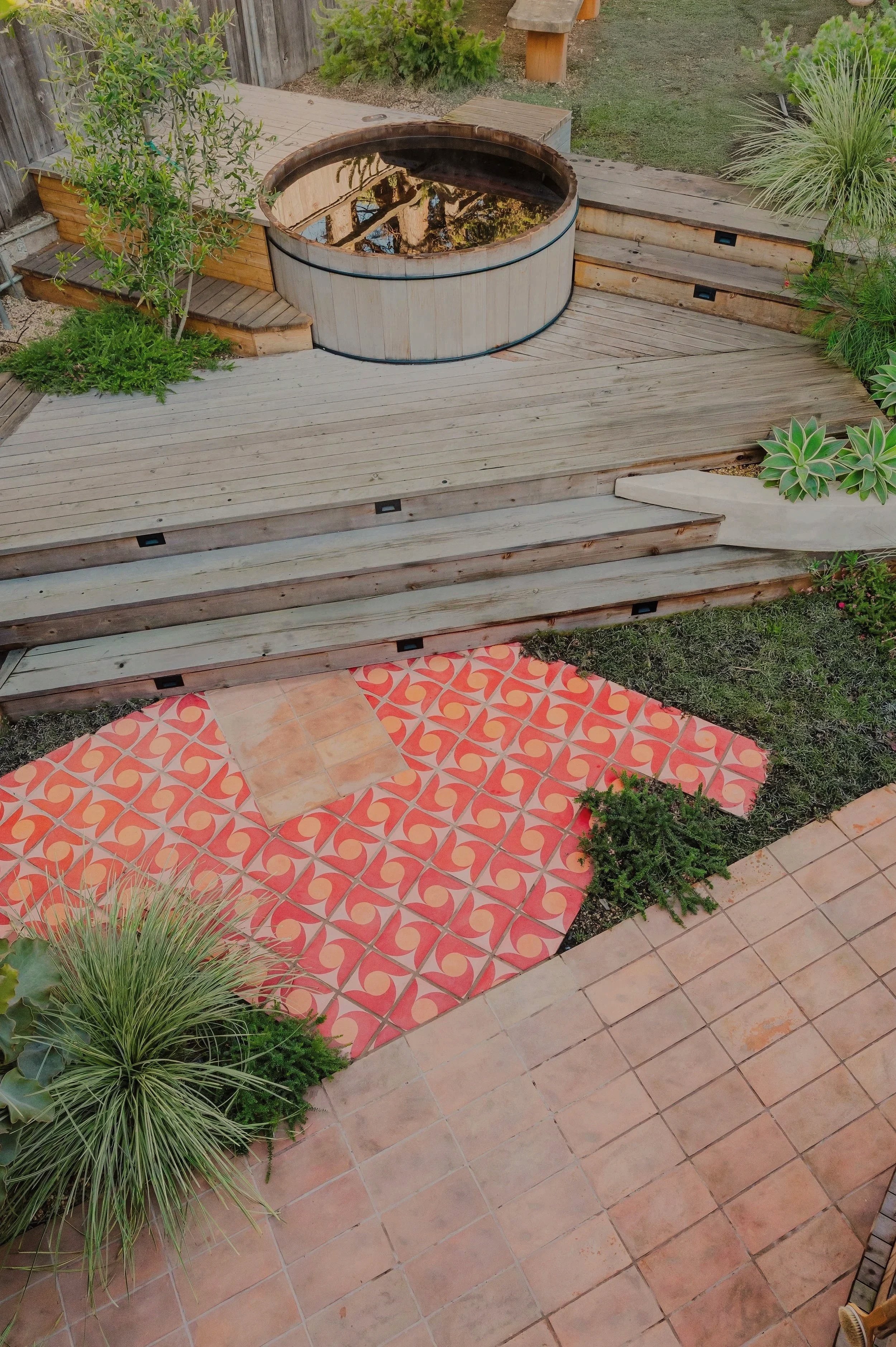 A backyard patio with a wooden deck, a small hot tub, tiled steps, and a patterned red and orange tile area with surrounding plants.