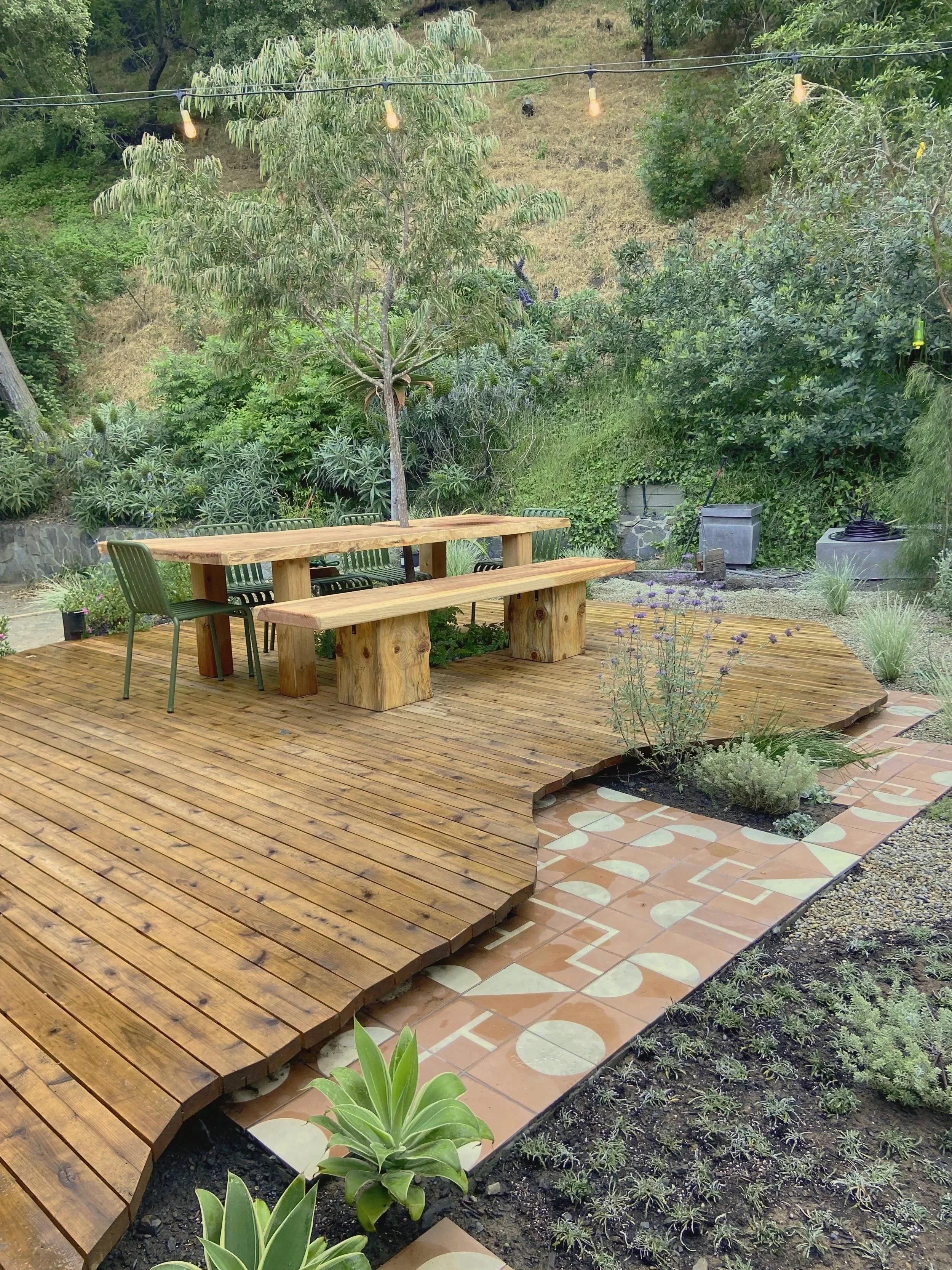 A landscaped backyard with a wooden deck and patterned tile pathway, surrounded by greenery and trees, with string lights hanging above.