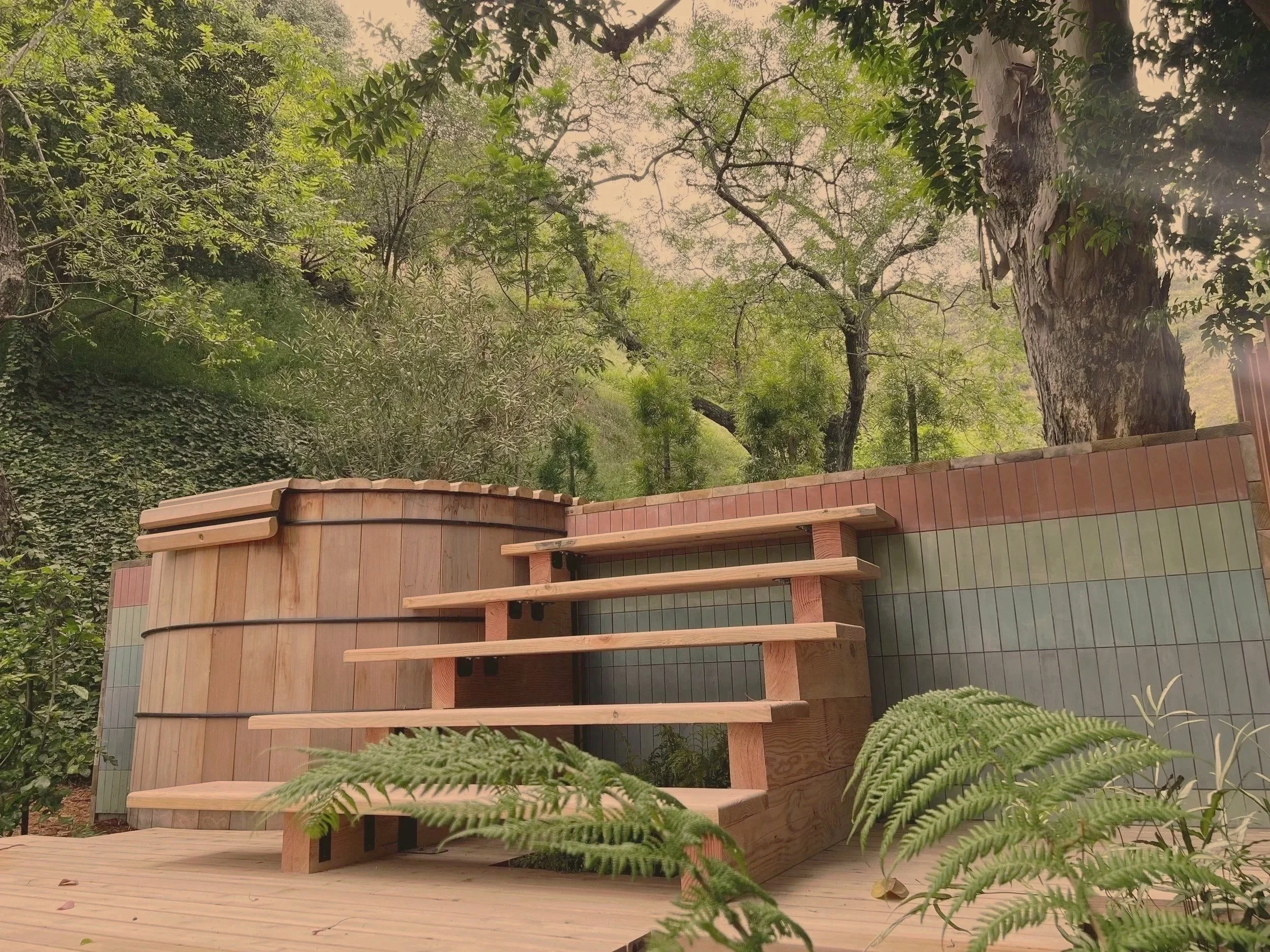 Wooden hot tub with deck and steps in a lush forest setting.