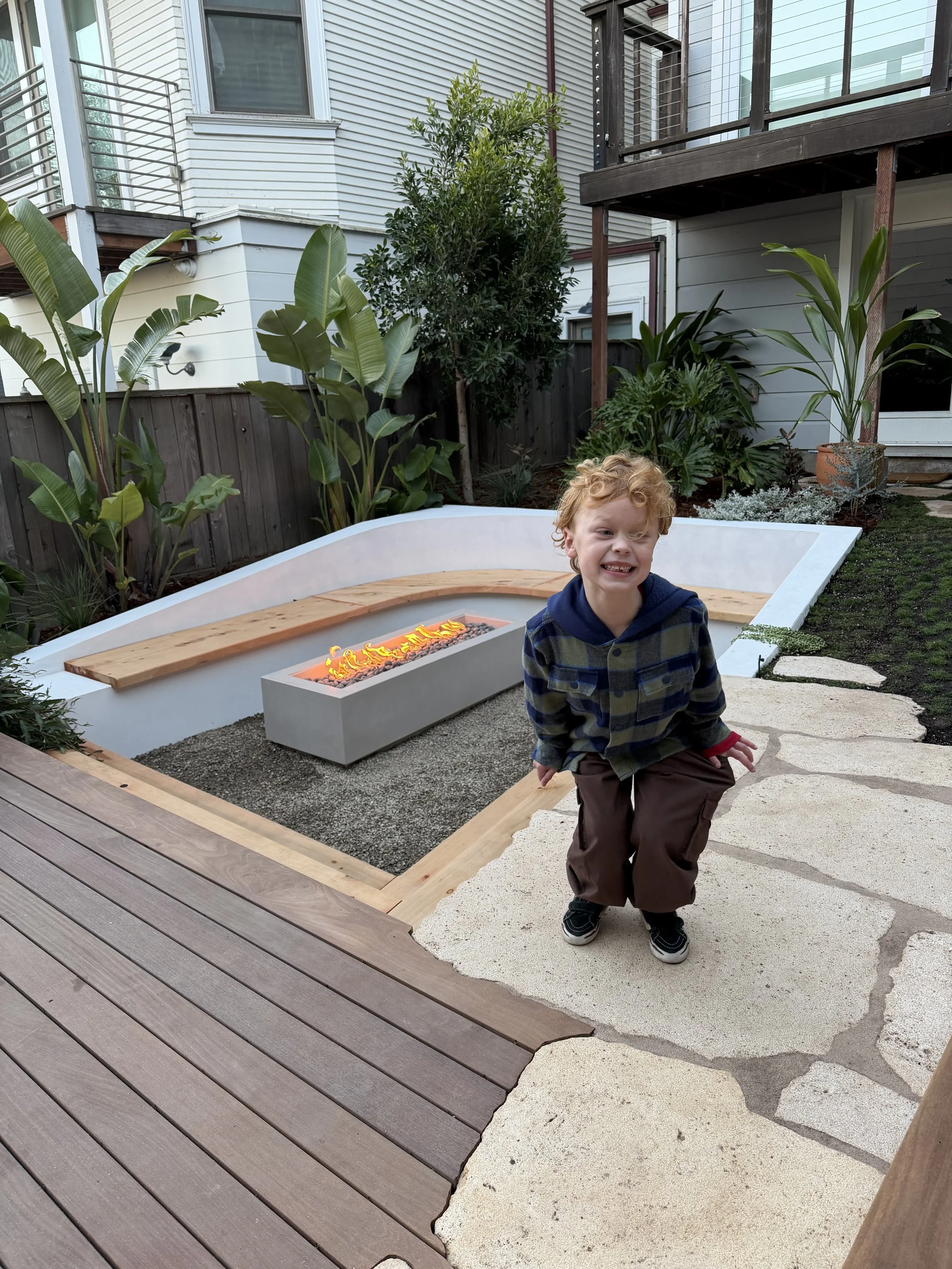 A child with curly red hair smiling and crouching in a backyard patio area. The patio has a mix of wooden and stone flooring, greenery, a modern fire feature, and is surrounded by a white house with large windows and outdoor plants.