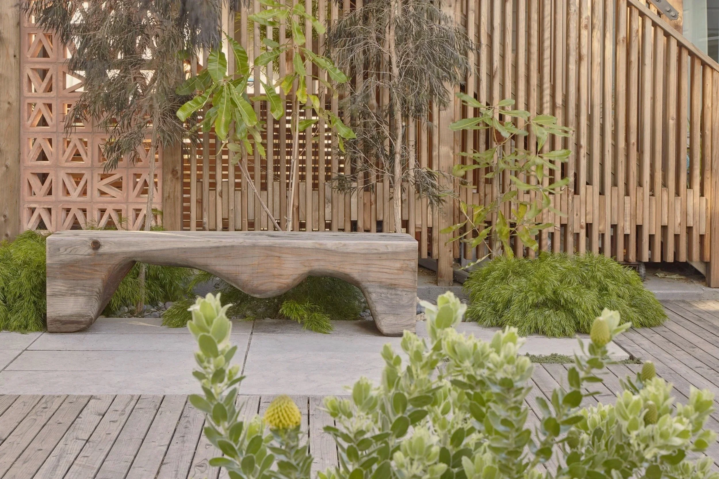 A wooden outdoor bench placed on a patio surrounded by green plants and wooden fencing.