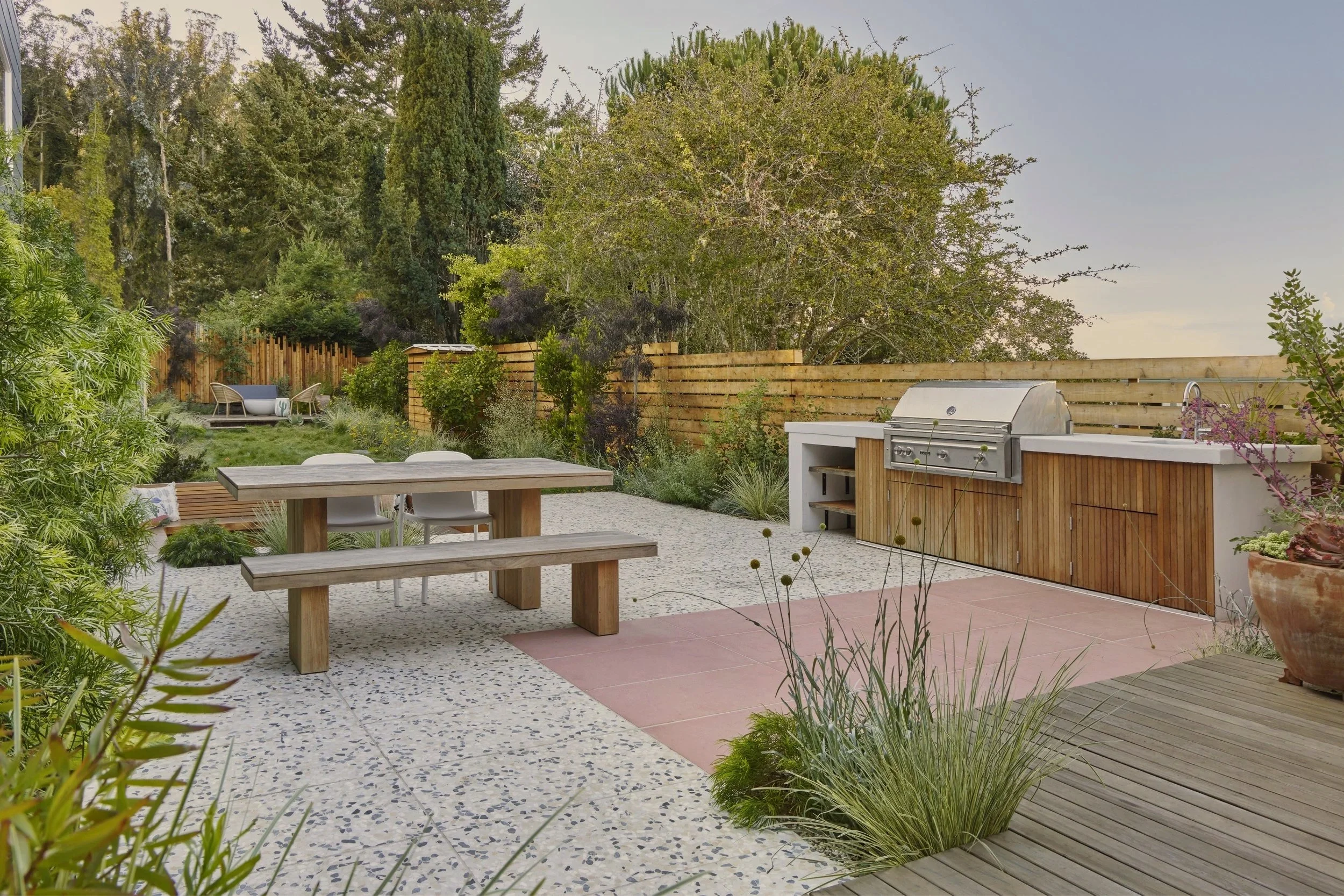 A backyard patio with a wooden picnic table and chairs, a built-in outdoor grill and sink, surrounded by various plants, bushes, and trees, enclosed by a wooden fence.