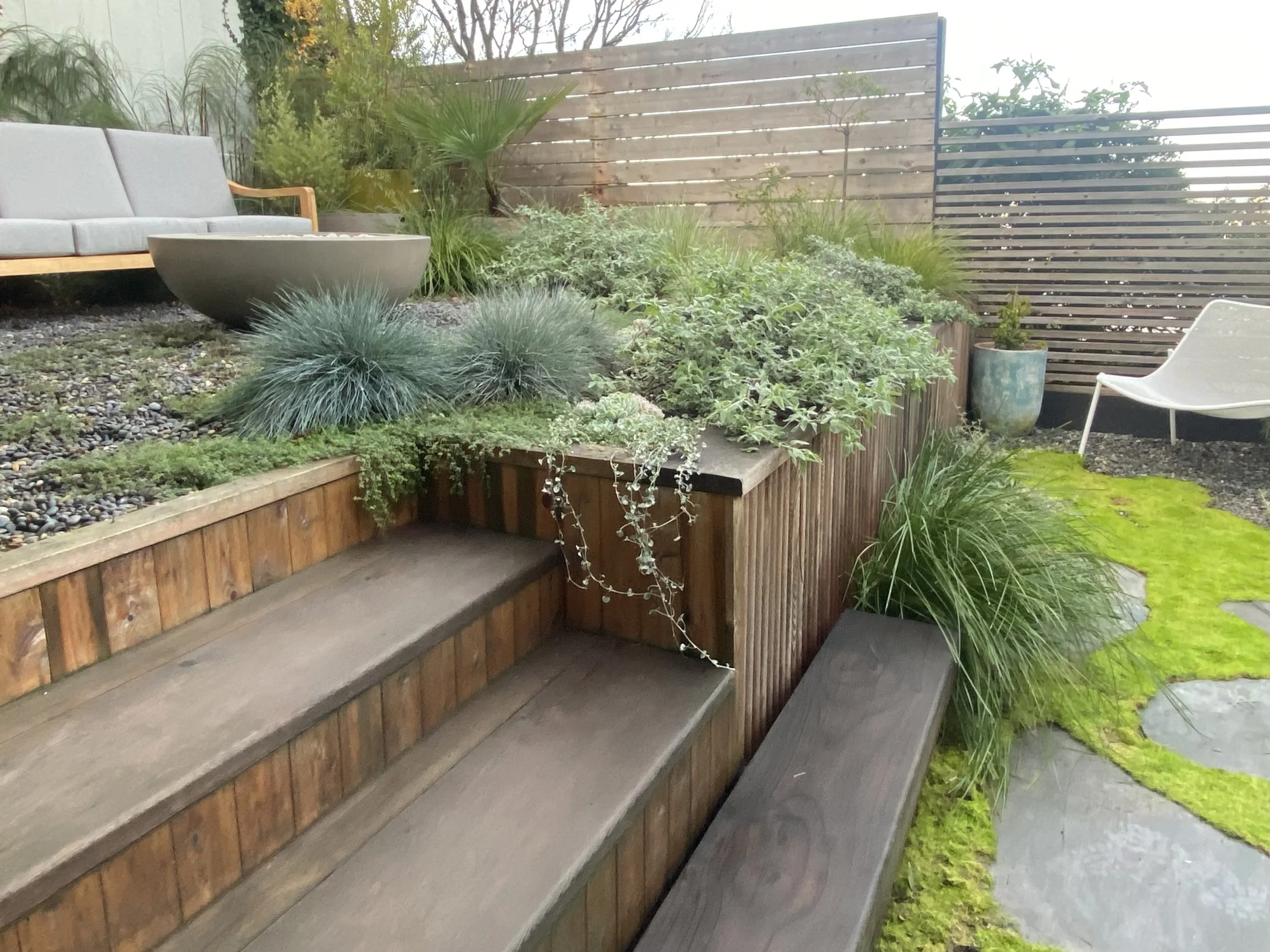 Outdoor garden with wooden stairs, a variety of green plants, a bench, and patio furniture.