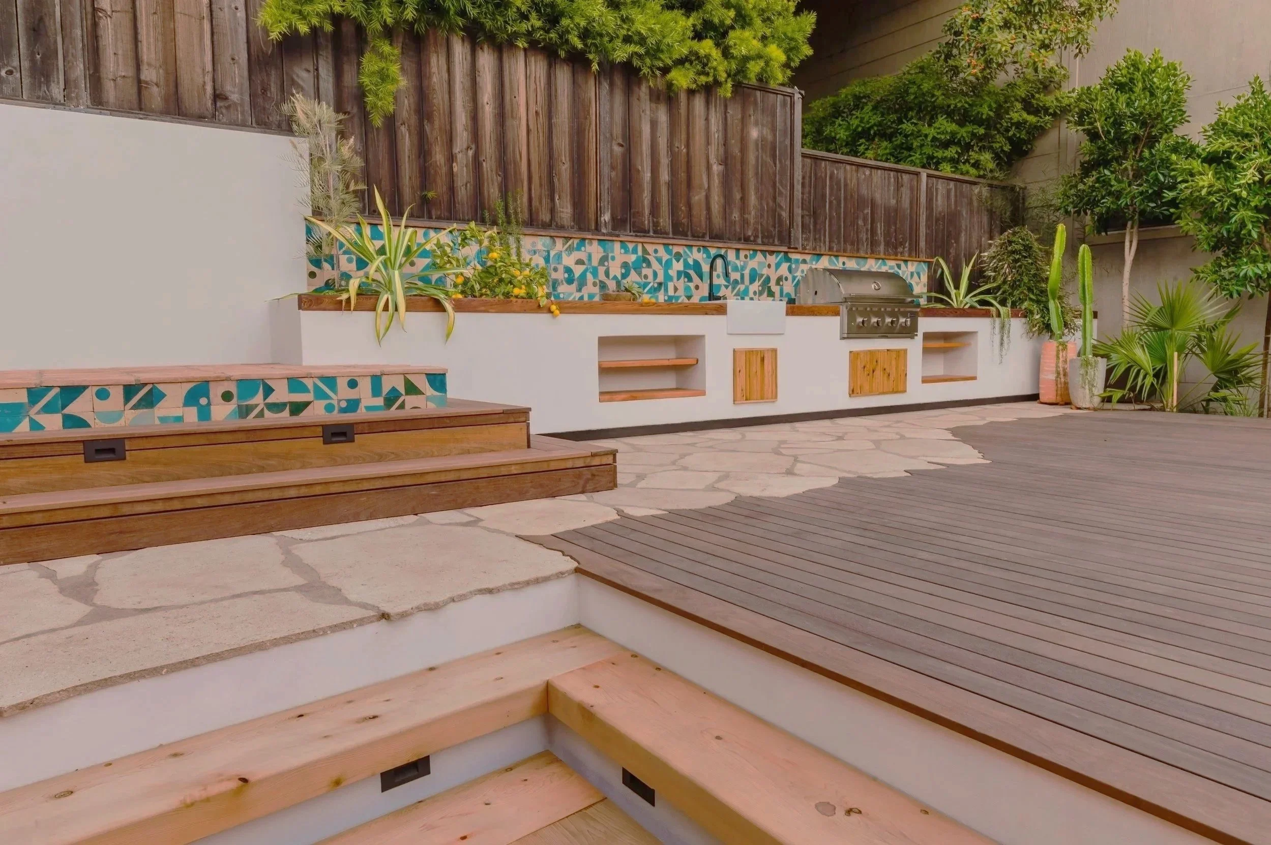 Backyard patio with wooden and stone steps, potted plants, a built-in grill area, and a decorative tiled wall, surrounded by a wooden privacy fence and lush green bushes.