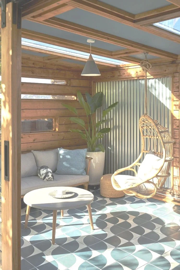 A sunlit outdoor patio with wooden walls and ceiling, featuring a beige sofa with pillows, a potted plant, a hanging rattan chair with cushions, a white side table, and patterned tile flooring.