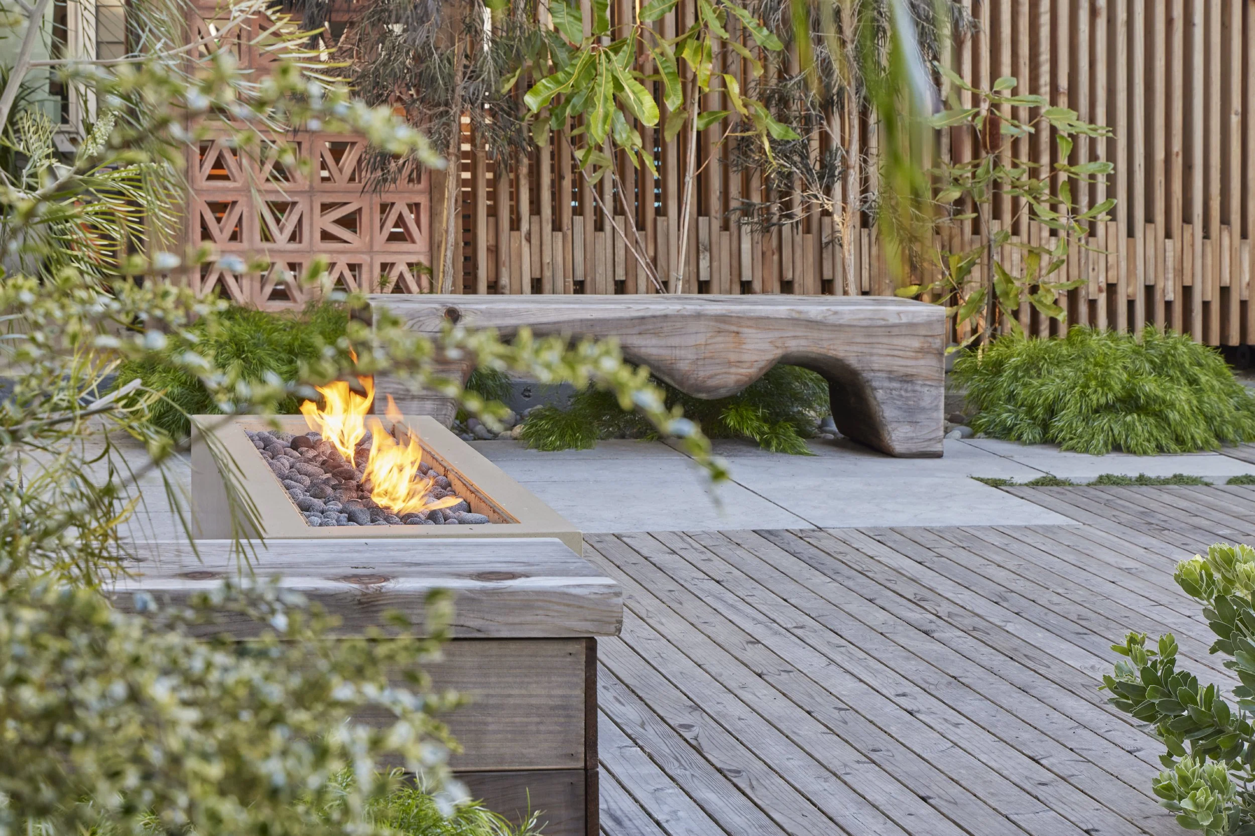 Backyard patio with a rectangular fire pit filled with dark stones and flames. There is a rustic wooden bench with an irregular shape behind the fire pit, surrounded by lush green plants. A wooden fence with a lattice pattern is in the background.