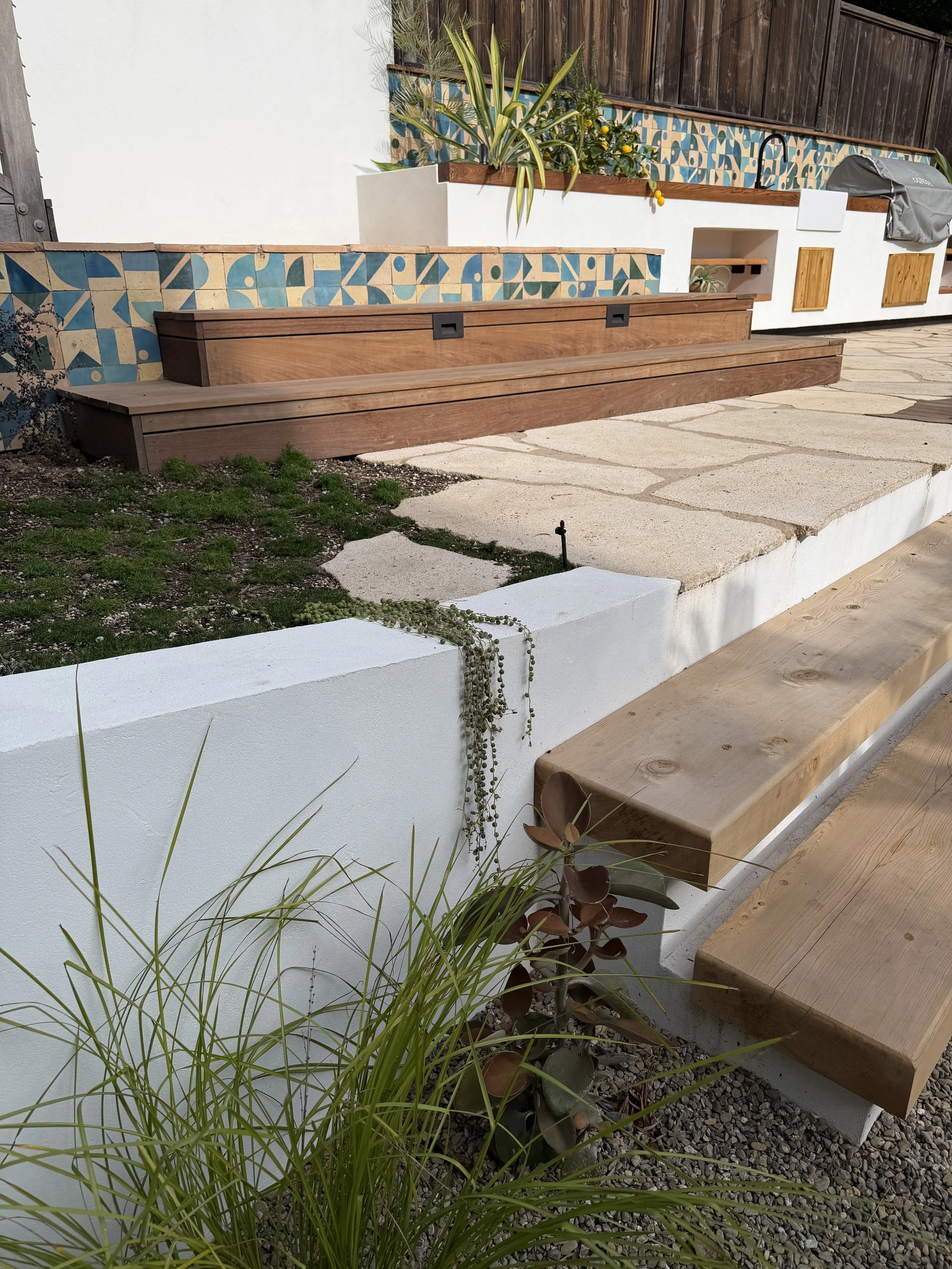 A backyard patio with a white concrete wall, wooden benches, potted plants, a tiled wall feature, stone steps, and greenery.