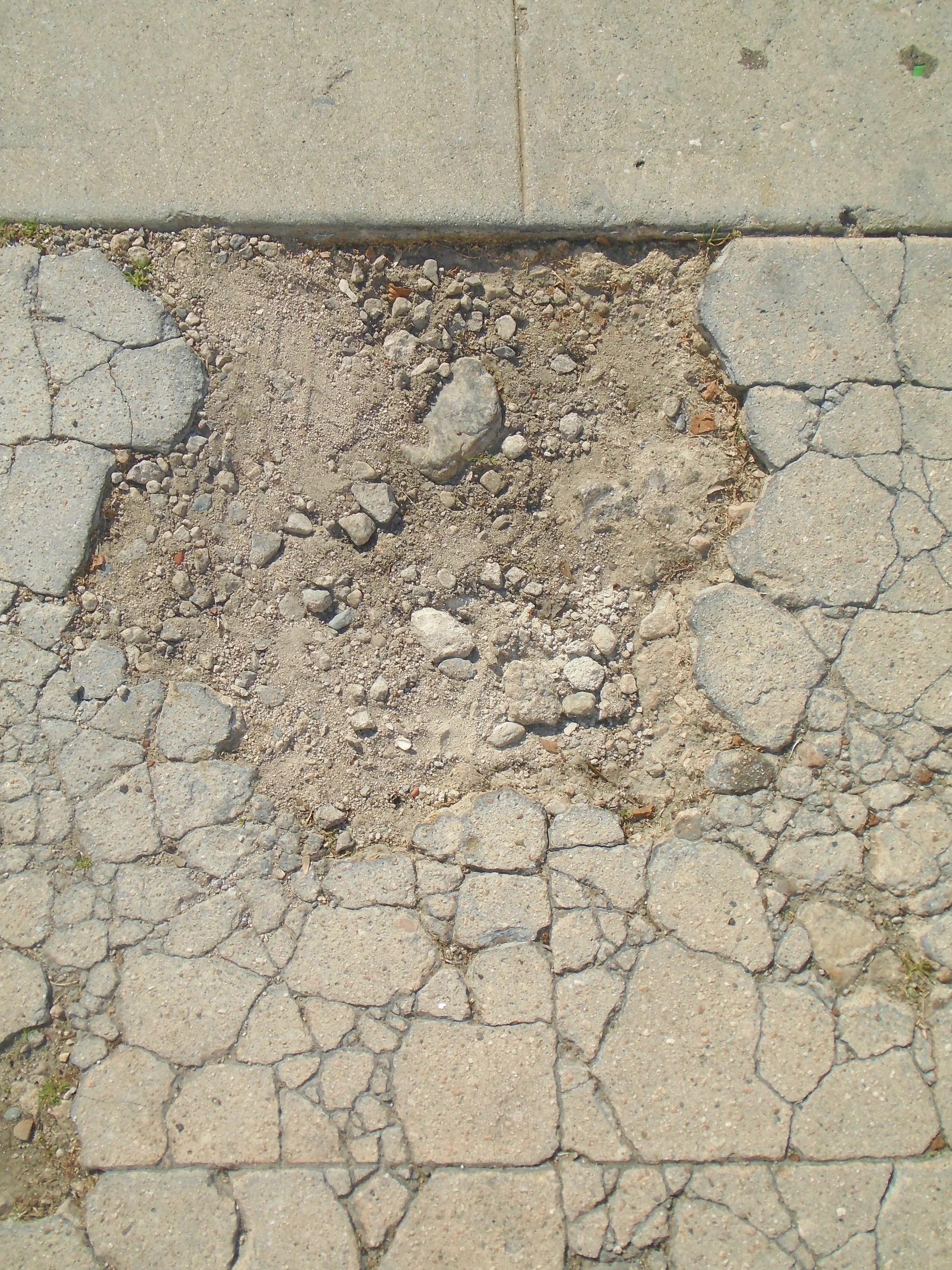 A section of broken and cracked concrete sidewalk with some dirt and small rocks visible.