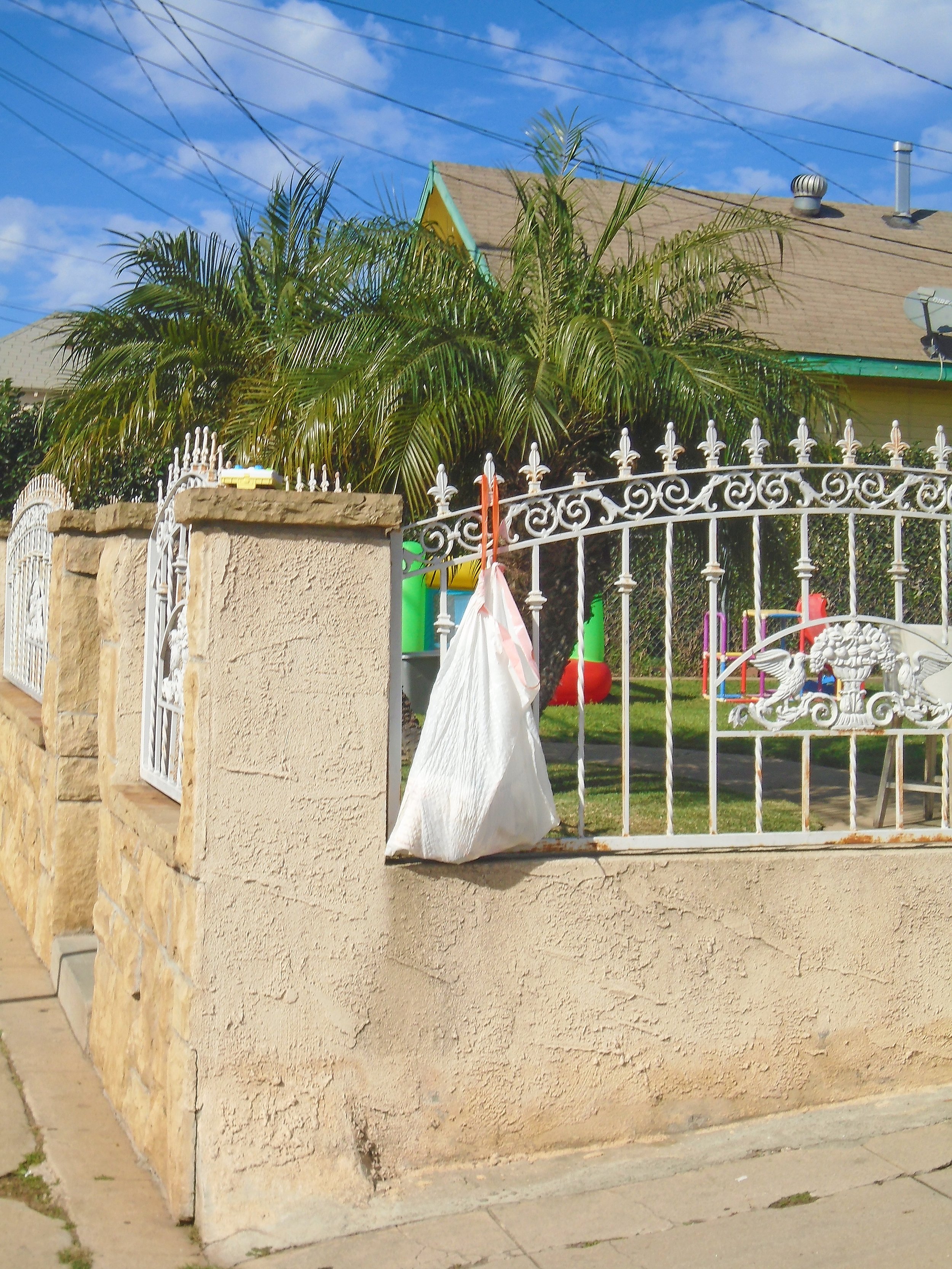 A residential front yard with a cream-colored stucco wall, an ornate white wrought iron gate, a palm tree, and children's toys visible in the background under a blue sky with wispy clouds.