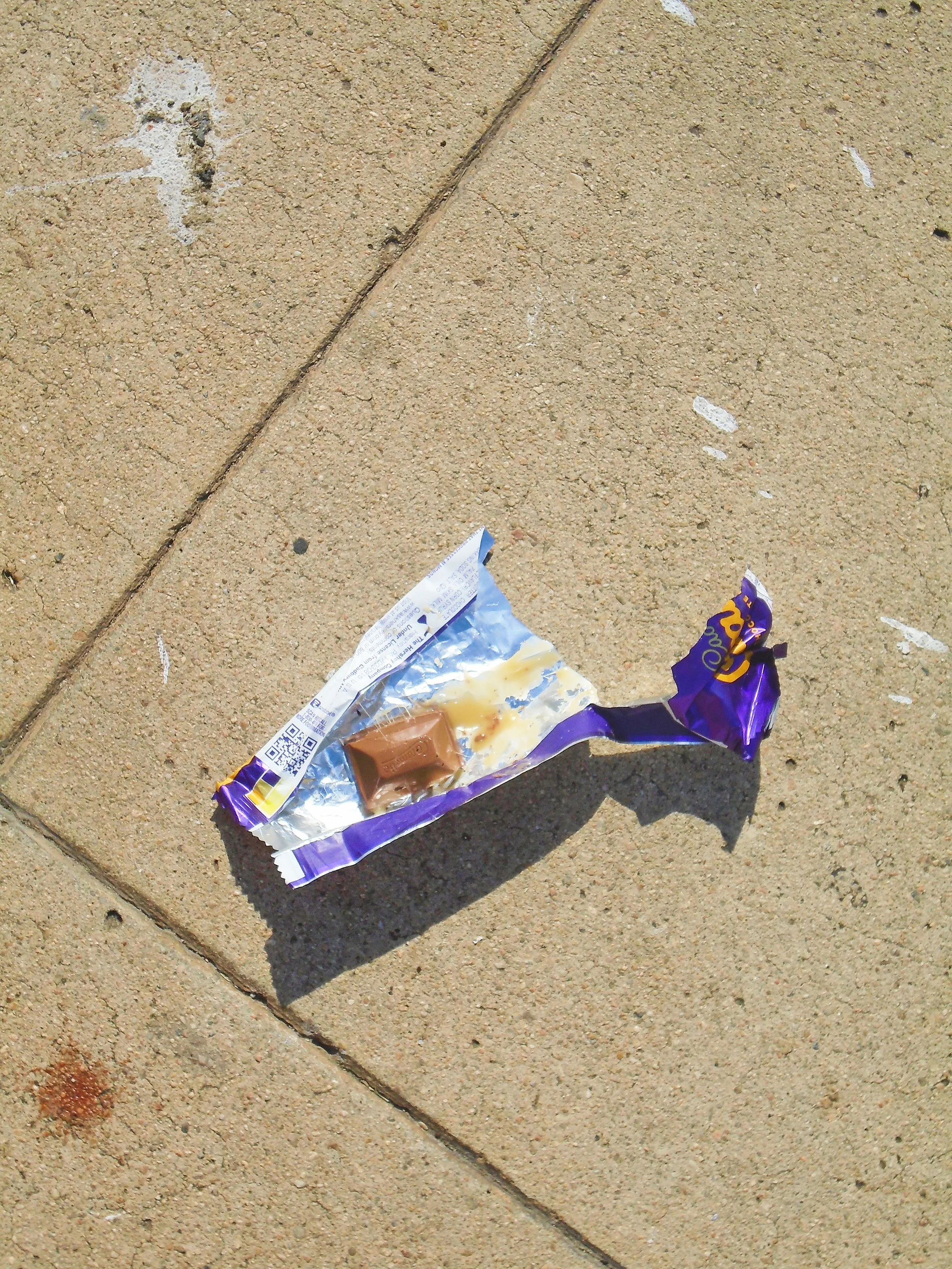 A discarded purple candy wrapper on a concrete sidewalk, with a piece of chocolate candy partially unwrapped.
