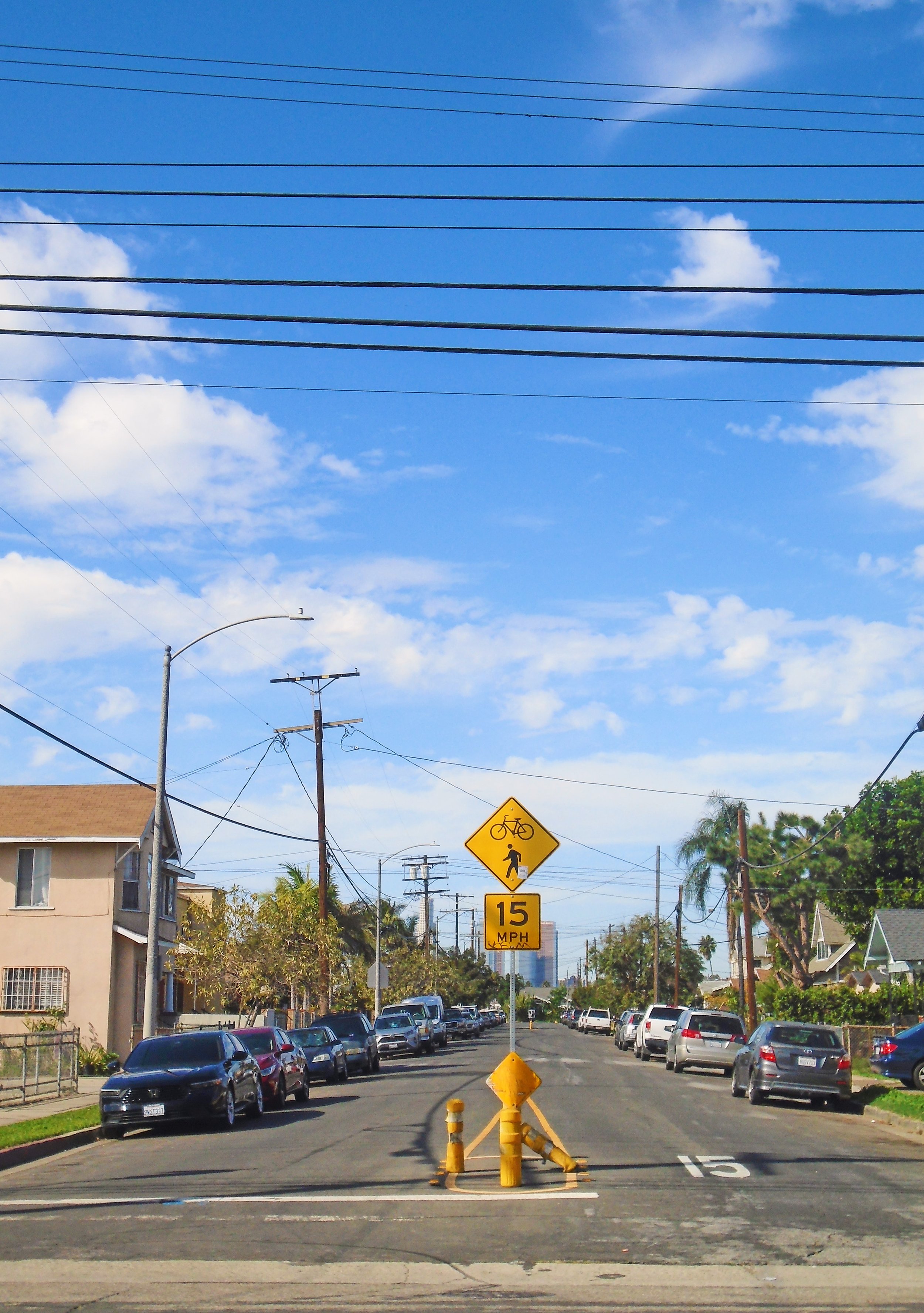 Street scene with parked cars, a yellow pedestrian and bike crossing sign, and a speed limit sign showing 15 MPH. Overhead, power lines stretch across a blue sky with some clouds.