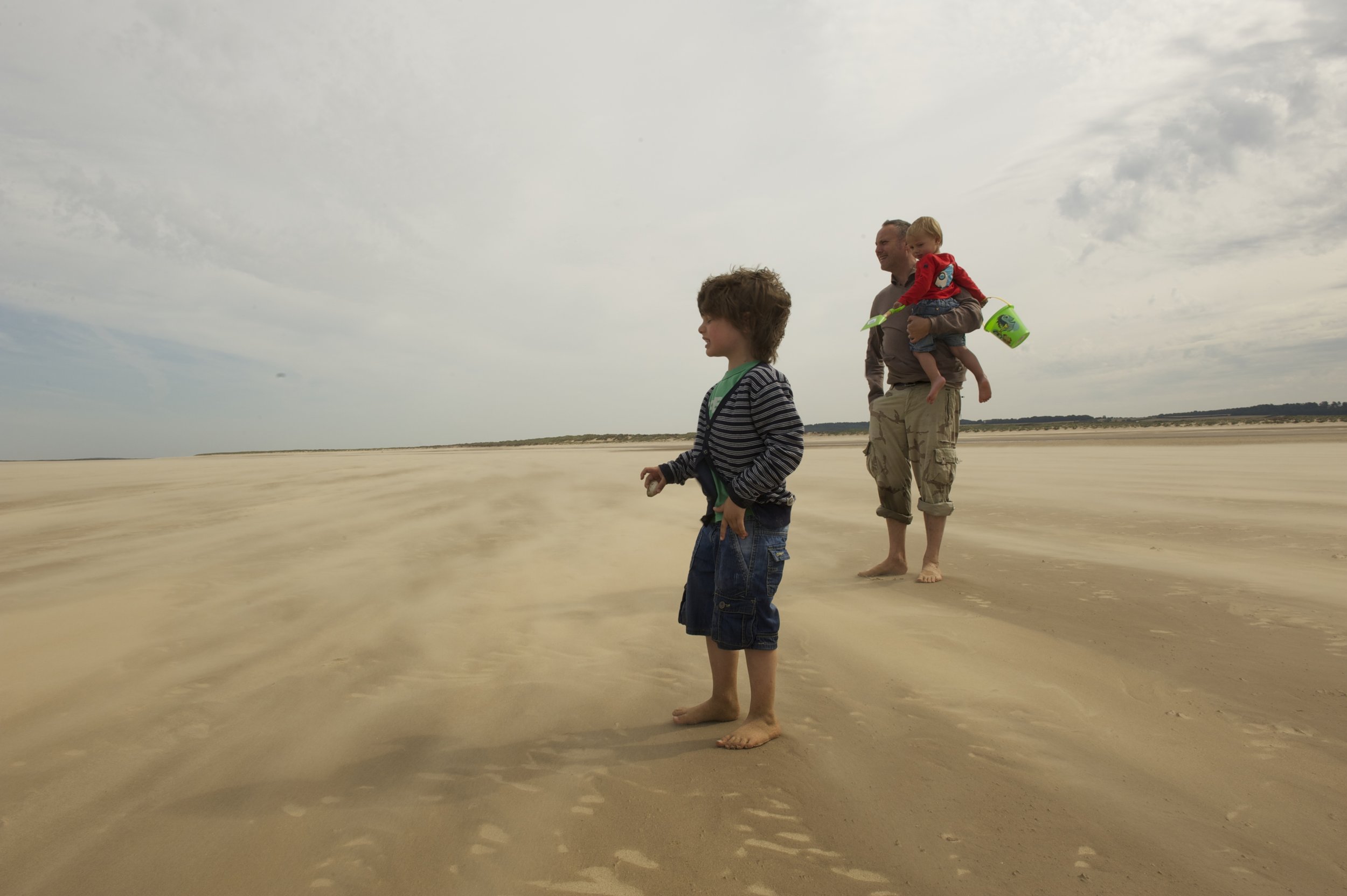 A father walking on the beach with two children, one older boy and a younger boy being carried, all barefoot on light sand under a cloudy sky.