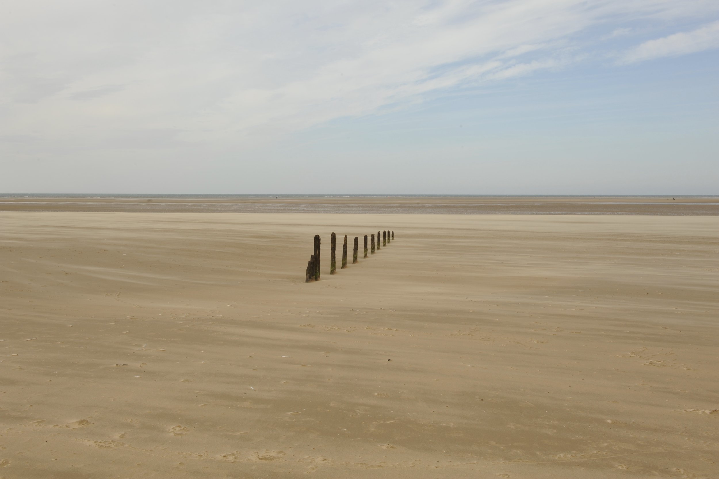 A wide sandy beach with a line of weathered wooden posts stretching into the distance under a partly cloudy sky.