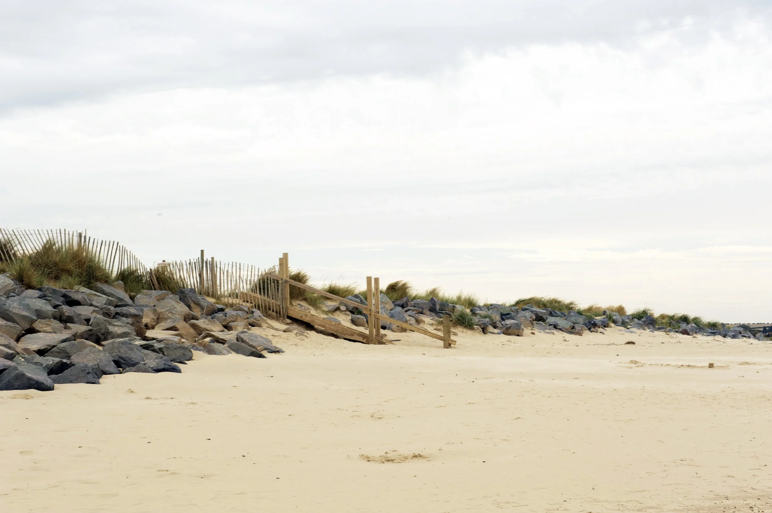 Empty sandy beach with a wooden staircase leading up a sand dune, rocks, and sparse grass, under a cloudy sky.