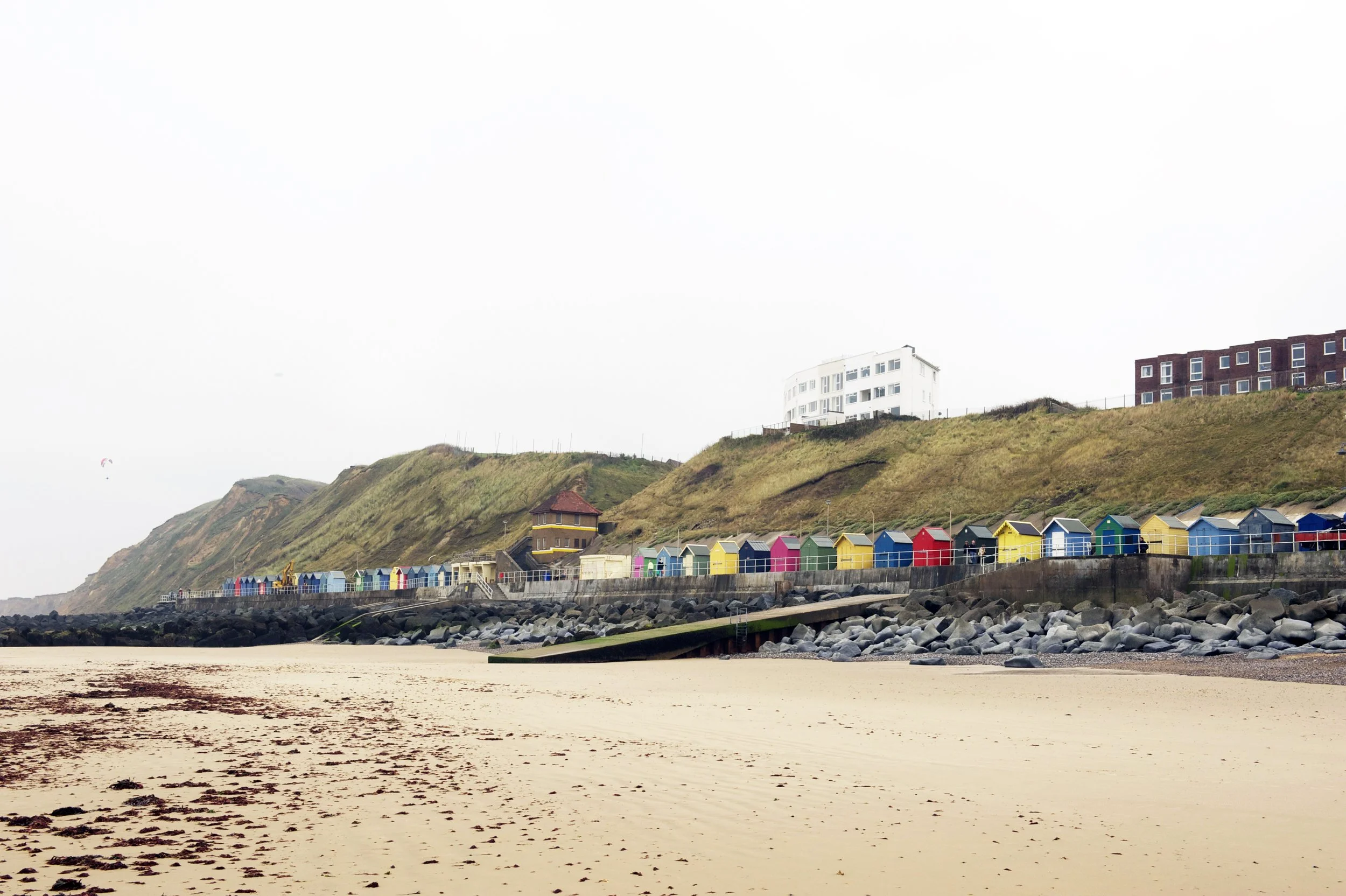 Beach with colorful beach huts along a rocky coastline, with grassy hills and buildings in the background under an overcast sky.