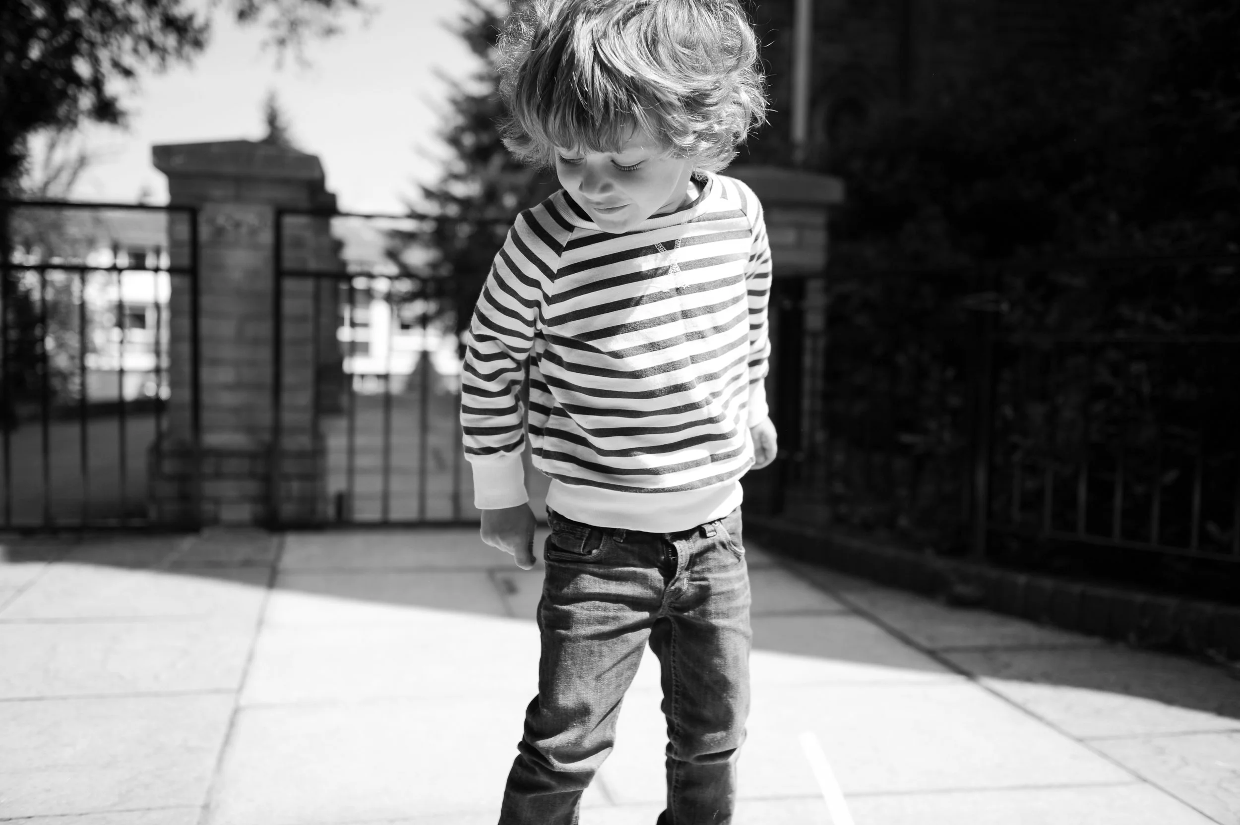 A young boy with curly hair wearing a striped long-sleeve shirt and jeans walking outdoors on a sidewalk.