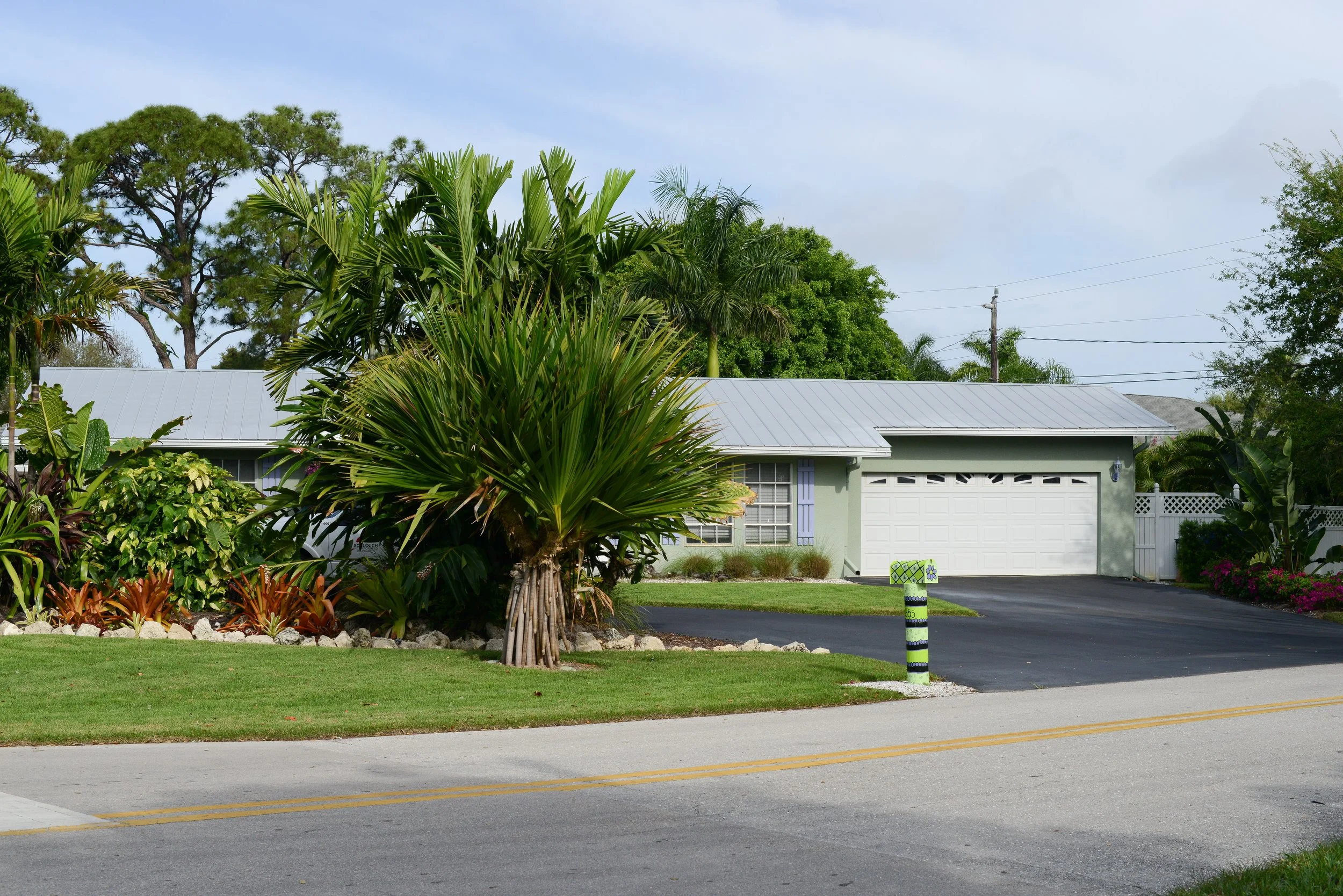 A house with a light green exterior and a white garage door, surrounded by tropical plants and trees, with a black asphalt driveway and a painted post near the street.