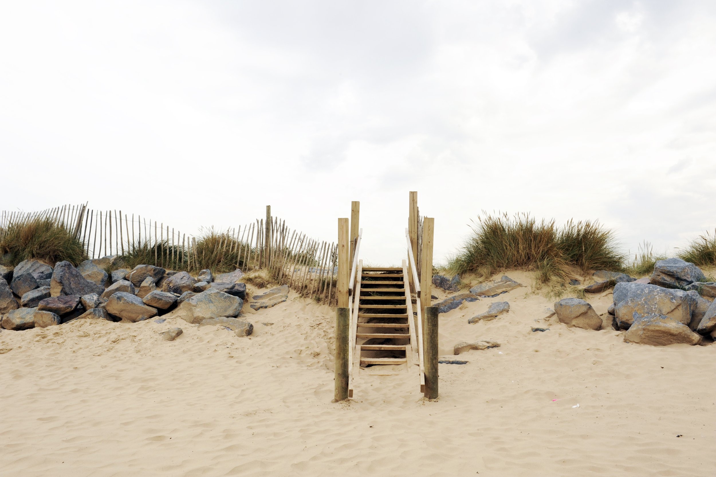 Wooden stairs leading up sandy beach from shoreline, flanked by rock formations and sea grass, with worn wooden fence on both sides and a cloudy sky overhead.