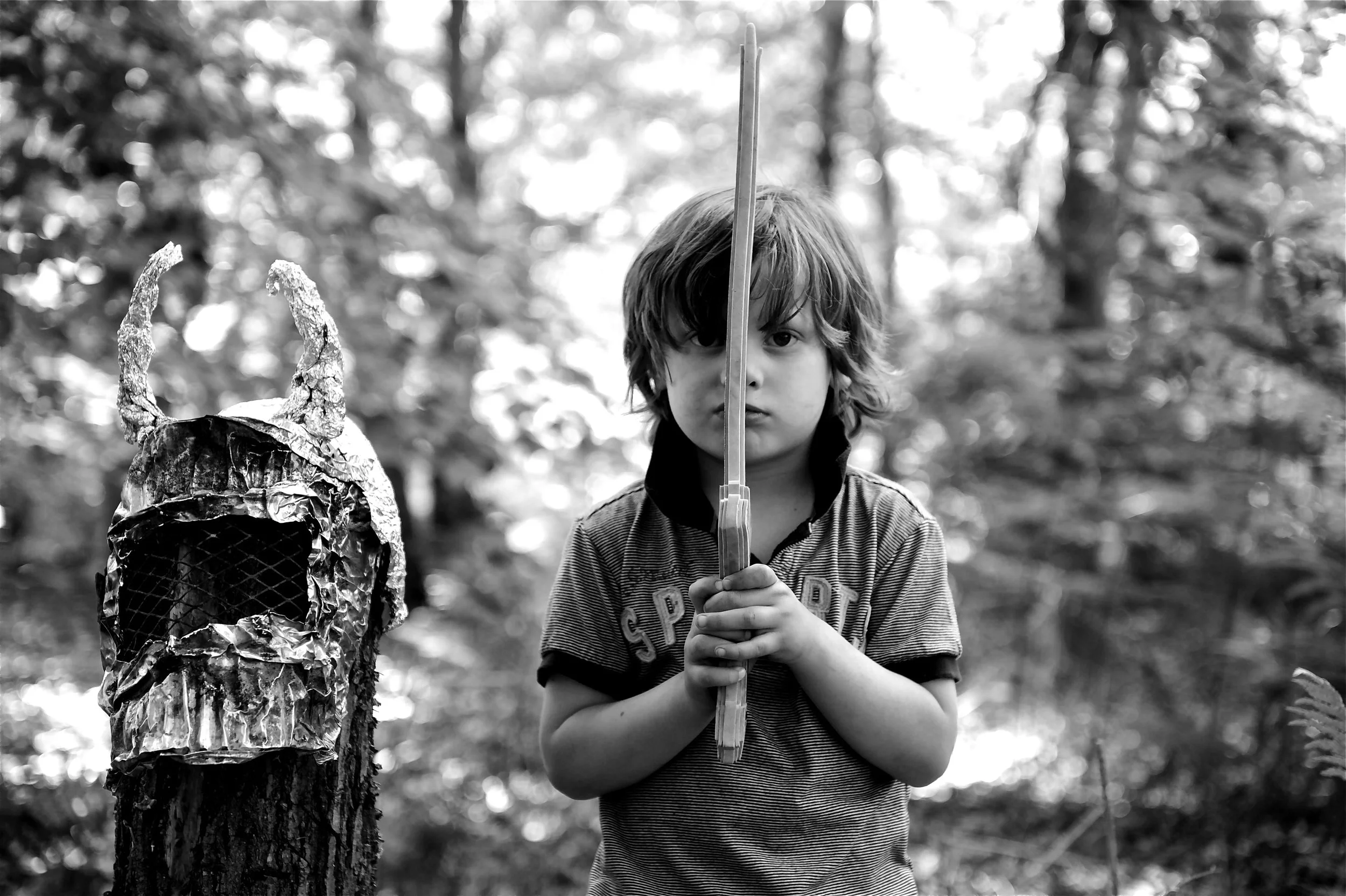 A young boy with shoulder-length hair, holding a sword upright with both hands, standing outdoors in a wooded area. There's a skull decorated with foil and tape on a tree to his left.