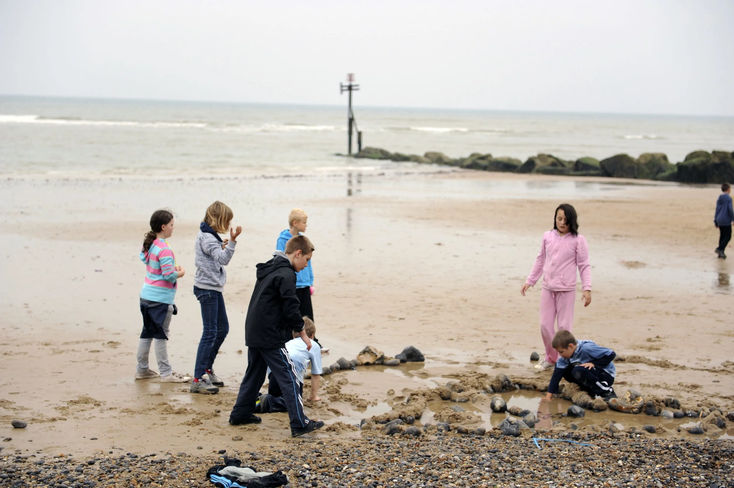 Children playing on a sandy beach with rocks and shallow water, some children kneeling and others standing, with the ocean in the background and overcast sky.