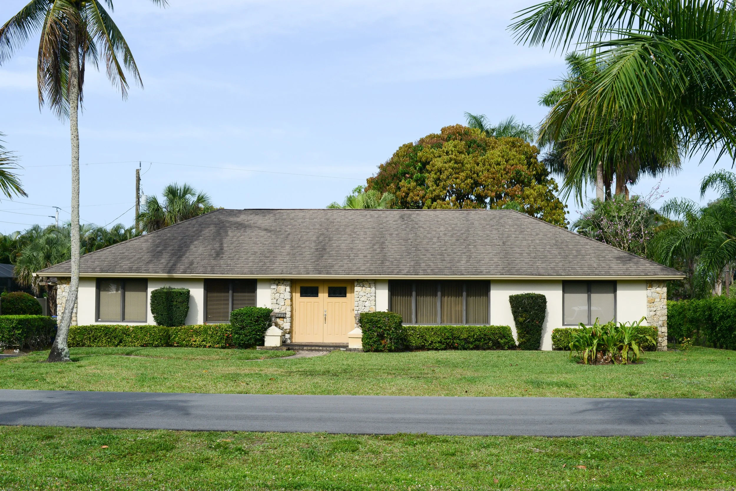 Single-story house with a brown roof, beige front door, and multiple windows, surrounded by green bushes and trees in a suburban setting.