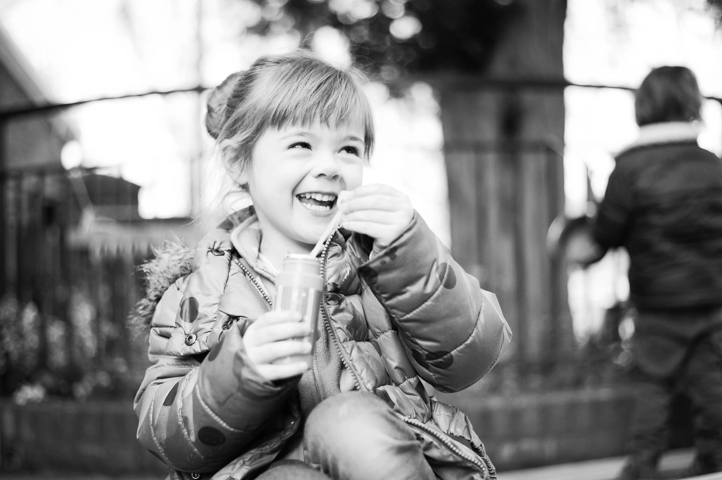 A young girl with a ponytail, wearing a jacket with polka dots, smiling while drinking from a small juice box with a straw. In the background, a person with short hair, wearing a dark jacket, is facing away.