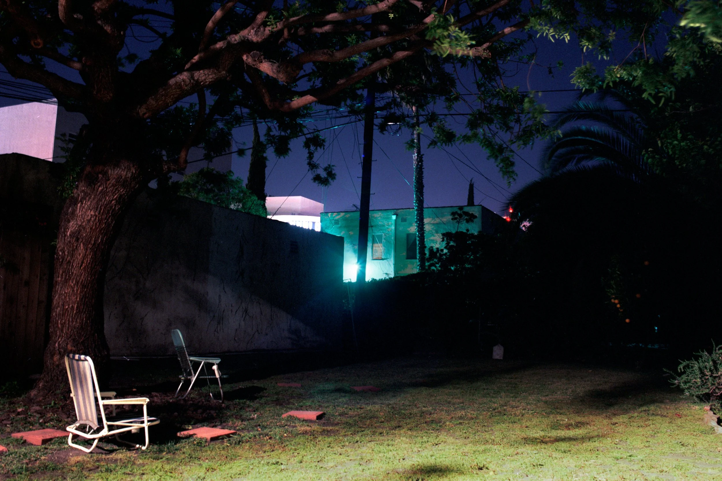 Nighttime backyard with two chairs on the grass, a large tree on the left, a concrete wall and illuminated houses and trees in the background.