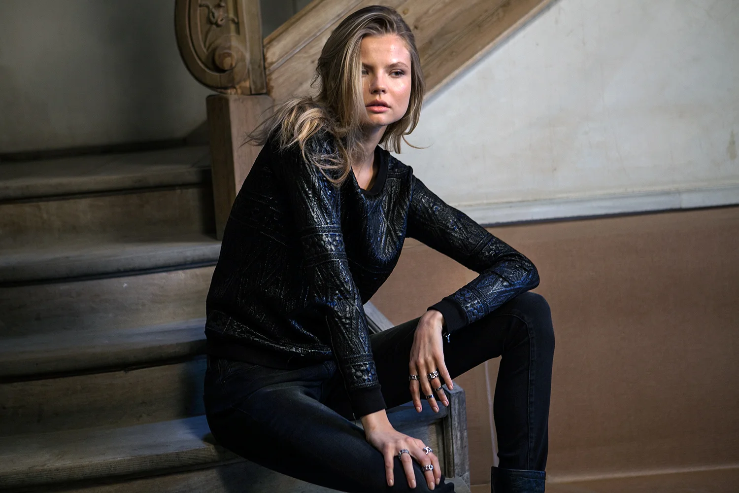 A young woman with blonde hair, wearing a black patterned jacket and black pants, sitting on wooden stairs indoors, with a serious expression and multiple rings on fingers.