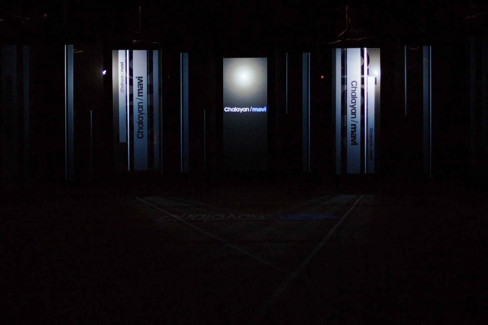 Dark room with illuminated signs displaying 'Chalayan/mavi' in the center and on the sides.