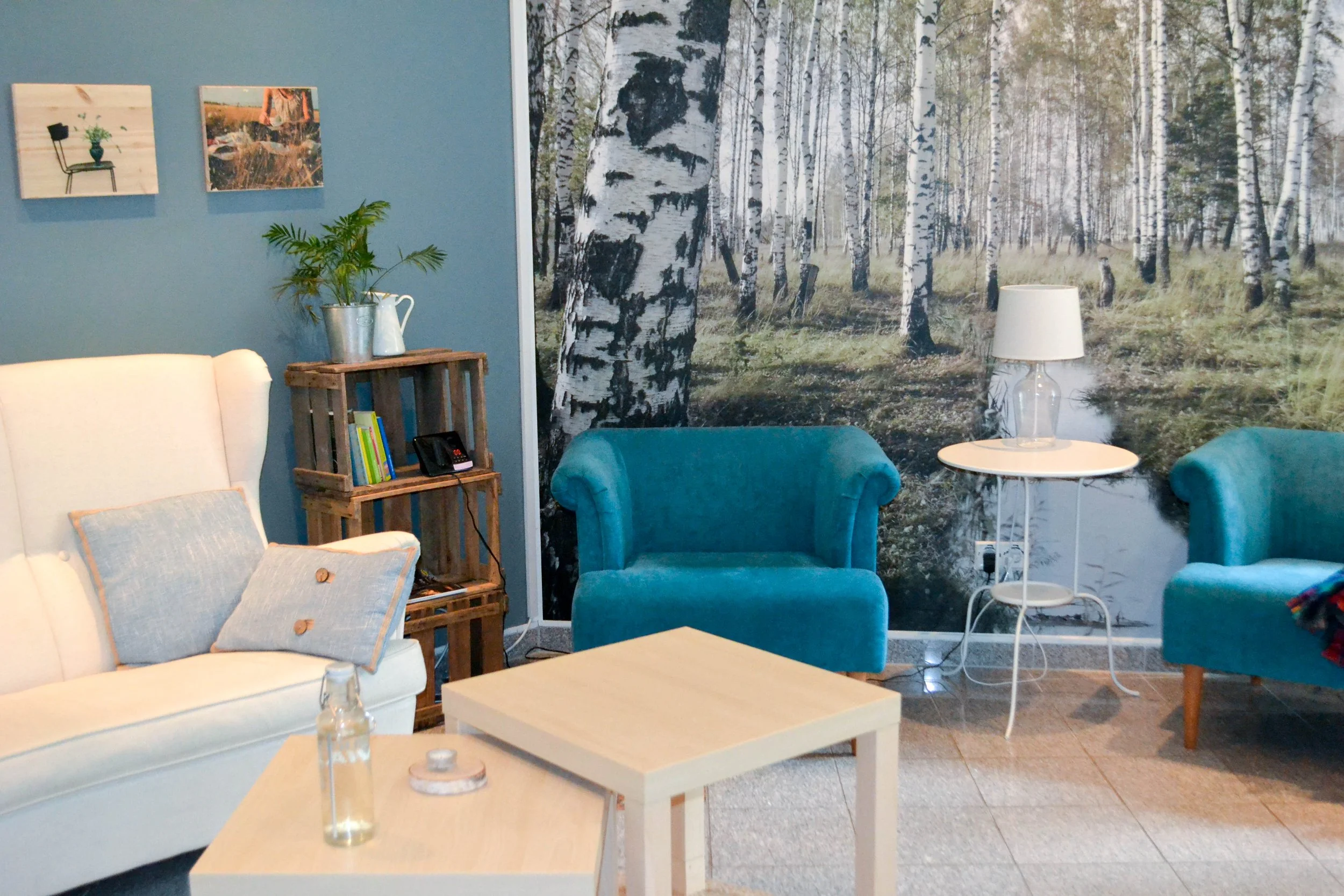 Ein Wohnzimmer mit blauer Wand und einer großen Waldtapete im Hintergrund, enthält eine weiße Couch mit Kissen, zwei blaue Sessel, einen kleinen runden Beistelltisch mit einer Lampe, einen quadratischen Couchtisch, eine Holzleiter mit Büchern und Dekorationsgegenständen, und eine Wanddekoration mit drei kleinen Fotos.