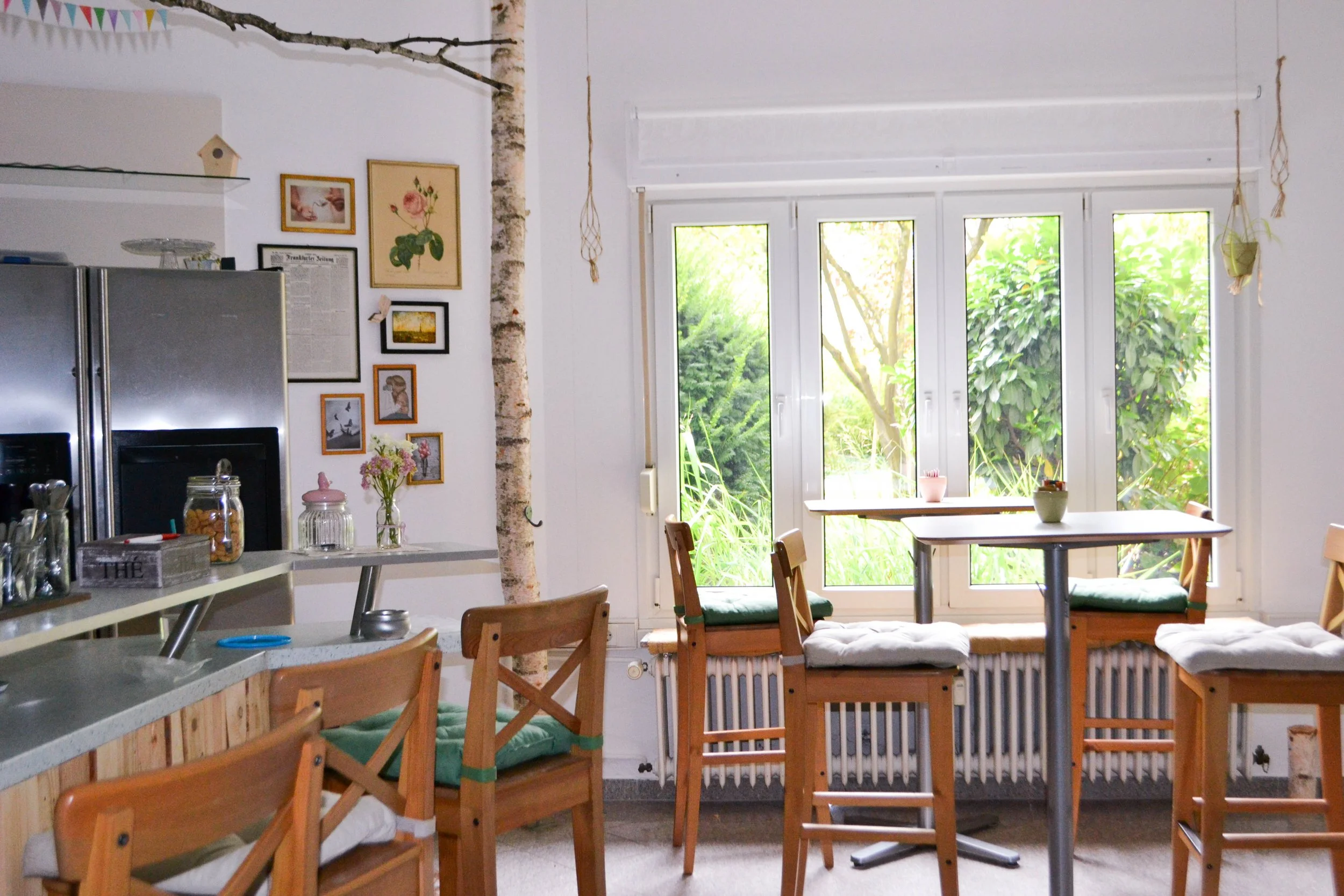 Gemütlicher Essbereich mit hoher Fensterfront, Holzstühlen mit Kissen, grünen Pflanzen im Garten, Wanddeko und eine Kücheninsel.